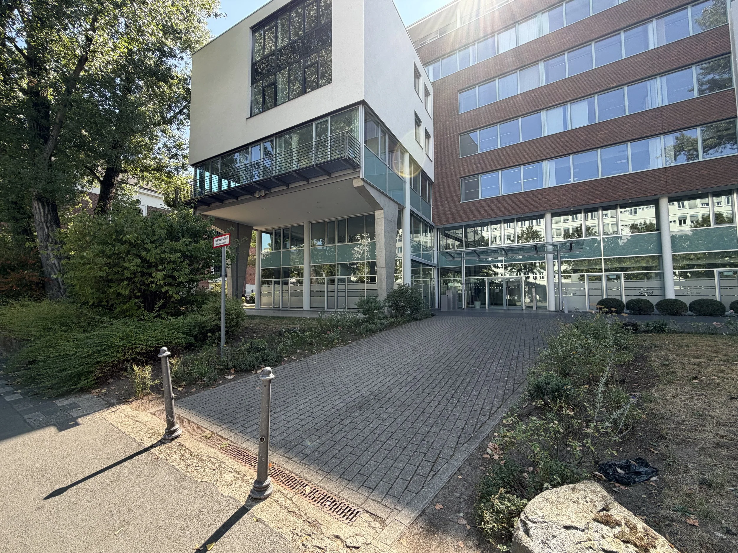 Mehrstöckiges Bürogebäude mit großen Fenstern und einem Balkon, umgeben von Bäumen und Grünflächen.
