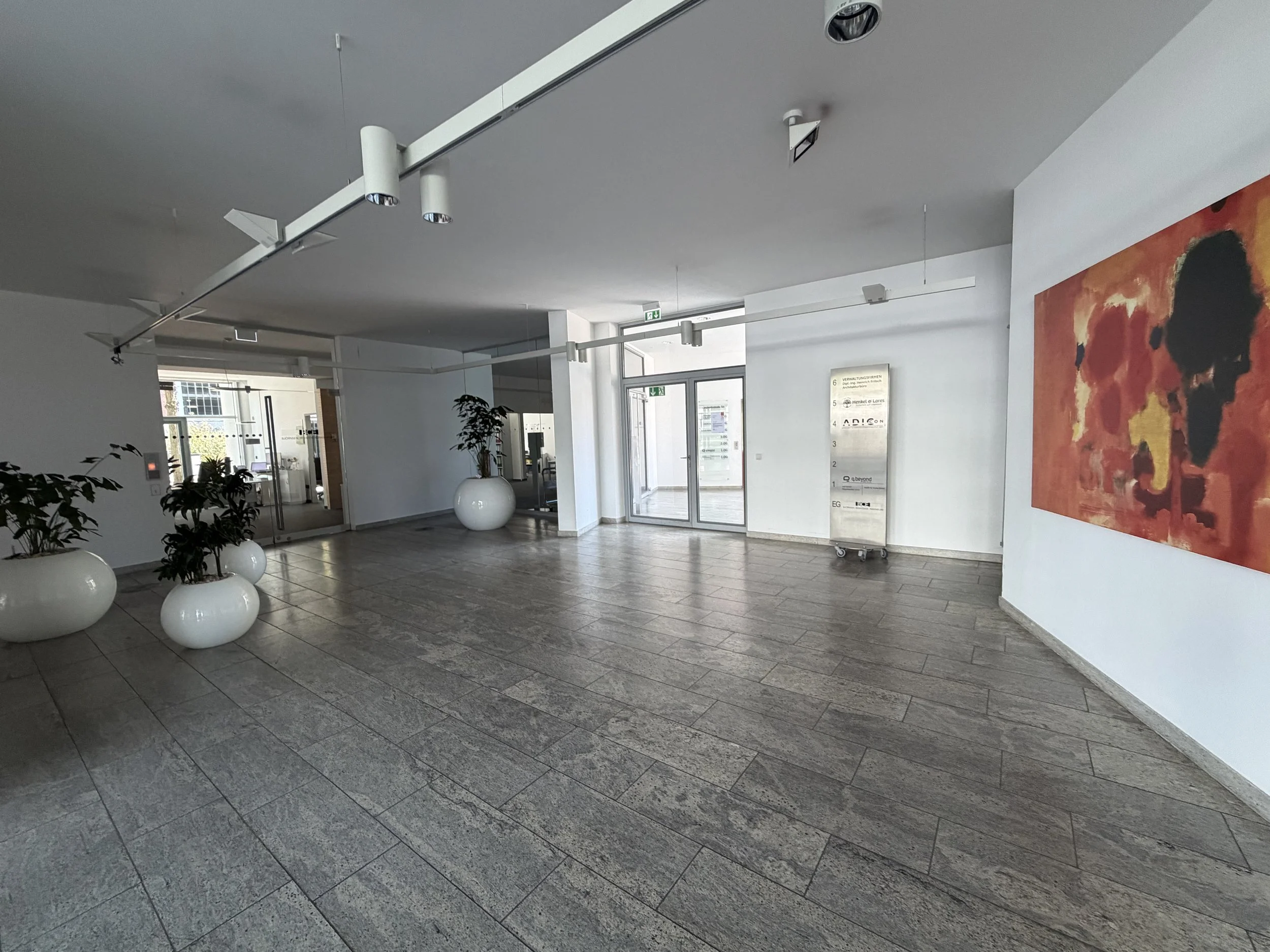 Ein heller Eingangsbereich mit grauen Fliesen, weißen Wänden und moderner Innenarchitektur. Es sind Pflanzen in weißen Töpfen, eine große bunte Kunstfotografie an der Wand, und ein Flur mit Glasschiebetüren sichtbar.