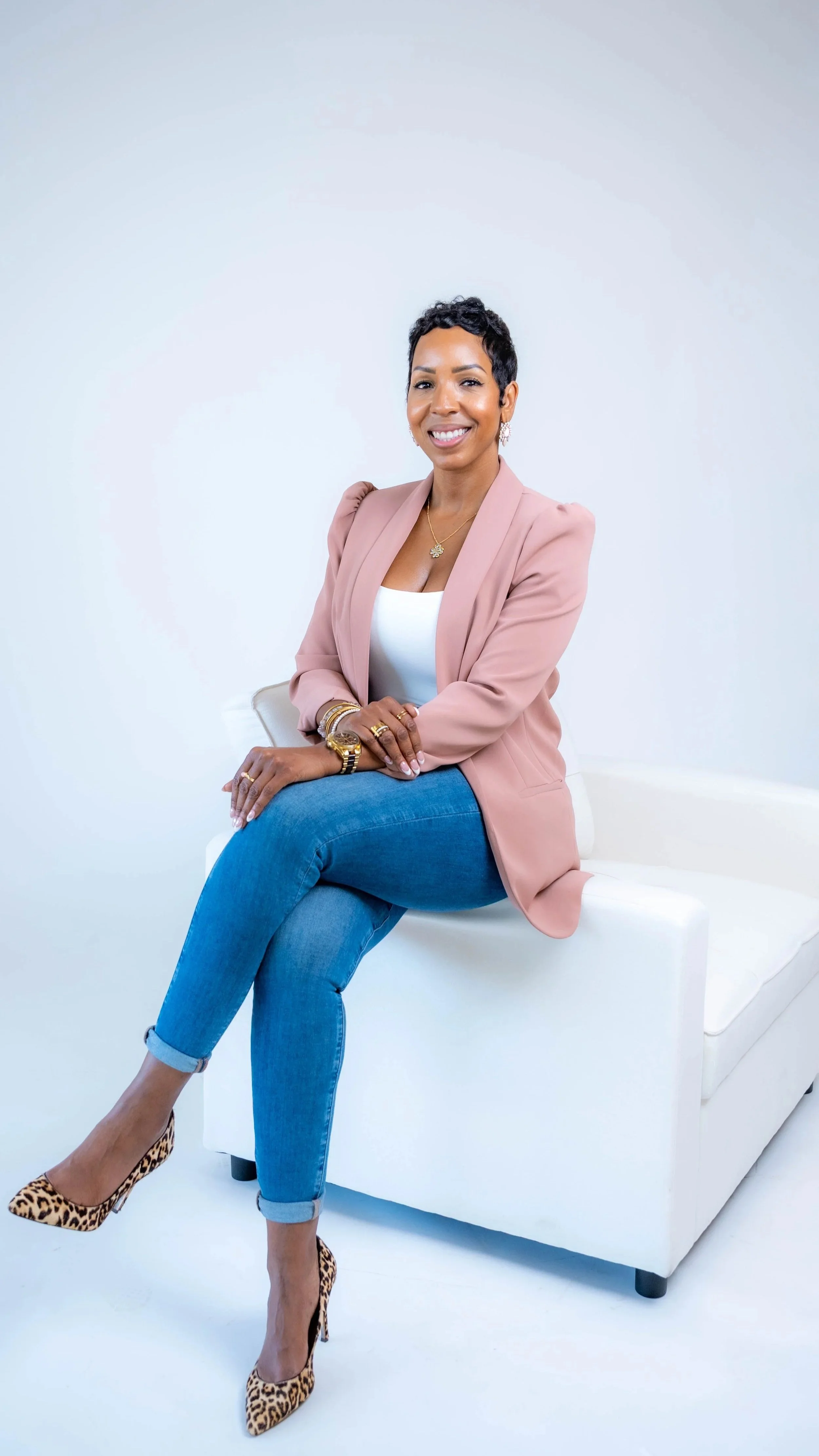 A woman with short black hair wearing a pink blazer, white top, blue jeans, and leopard print high heels sitting on a white armchair with a white background.