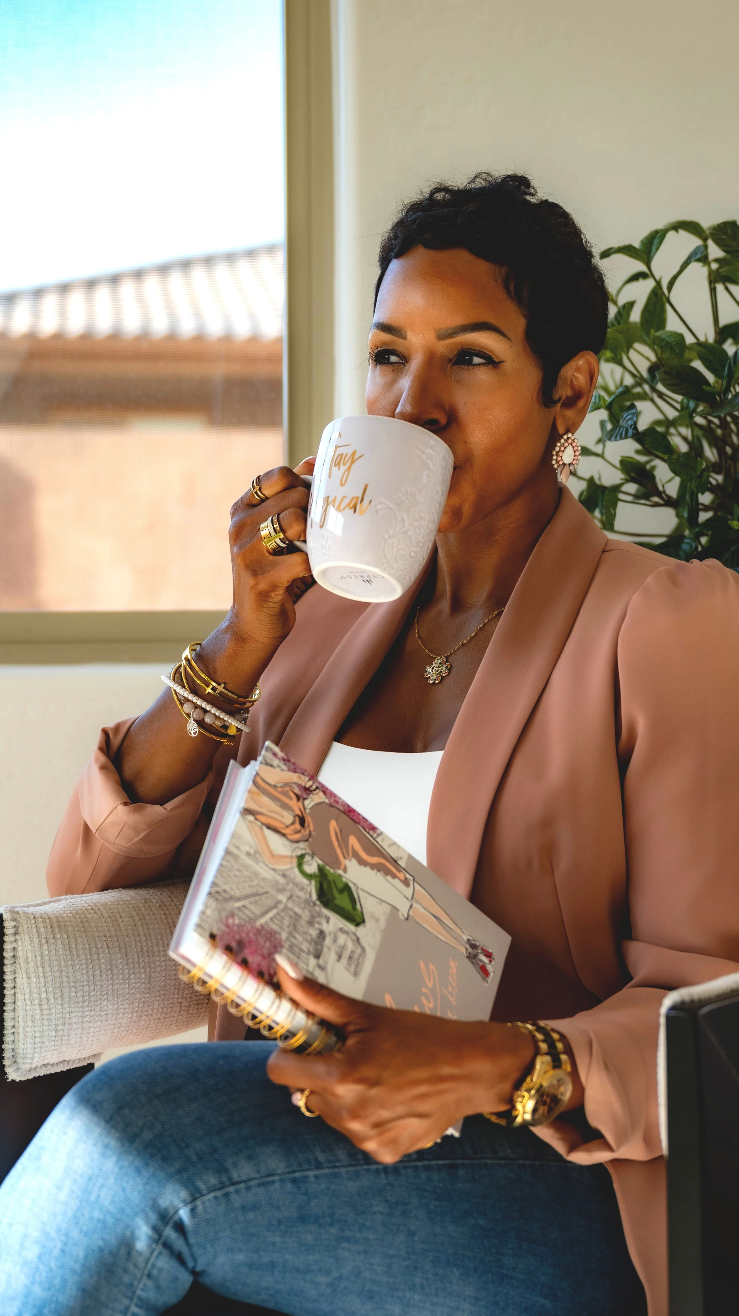 A woman with short black hair wearing jewelry, a beige blazer, and jeans, sitting indoors near a window, sipping from a mug that reads "Boss Lady" and holding a spiral-bound notebook with an illustration of a woman in a dress.