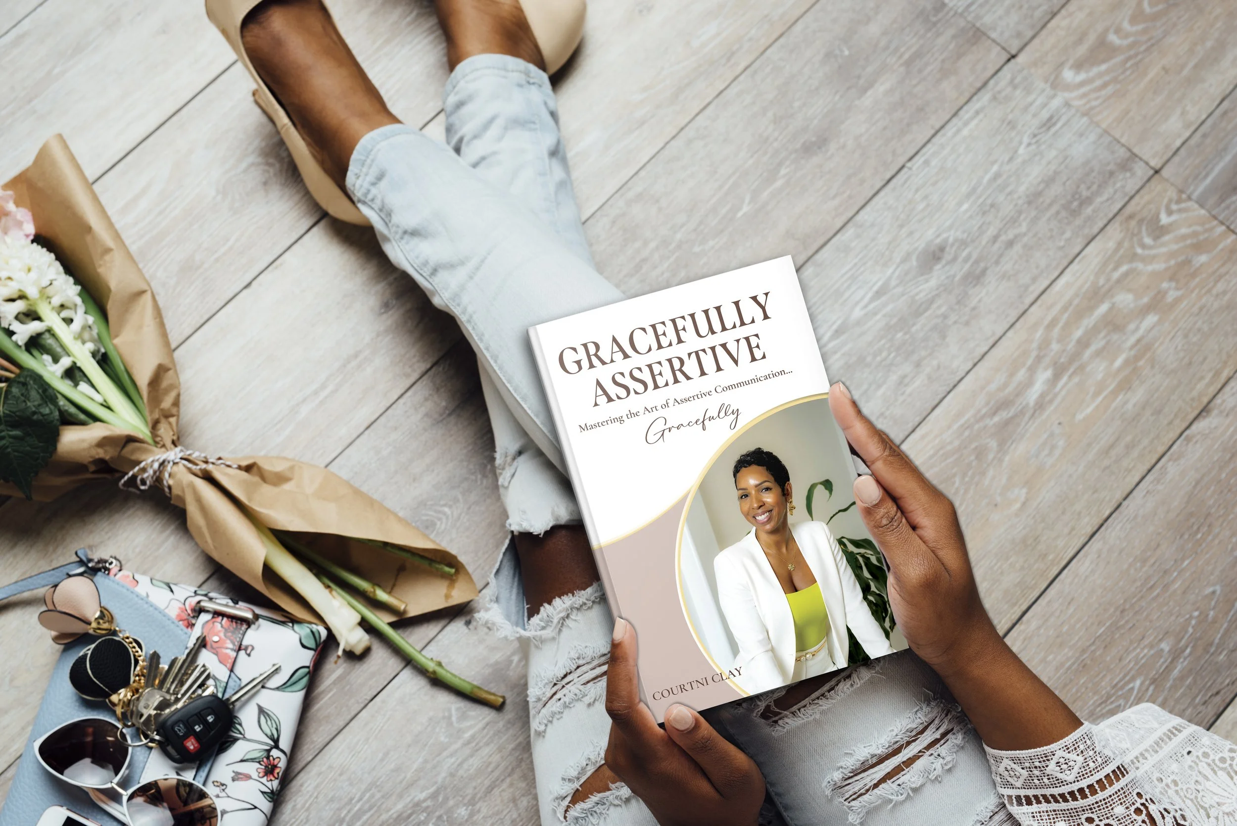A person sitting on the floor holding a book titled "Gracefully Assertive" by Courtni Clay, with flowers, sunglasses, keys, and a patterned bag nearby.