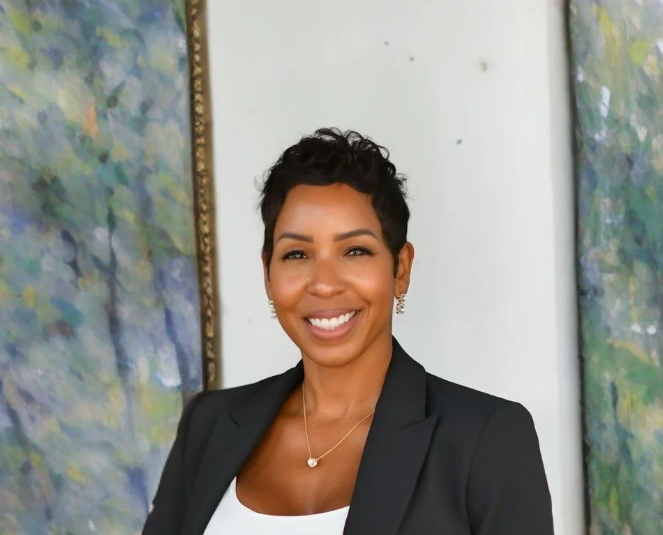 A smiling woman with short black hair, wearing a black blazer and jewelry, standing in front of abstract paintings.
