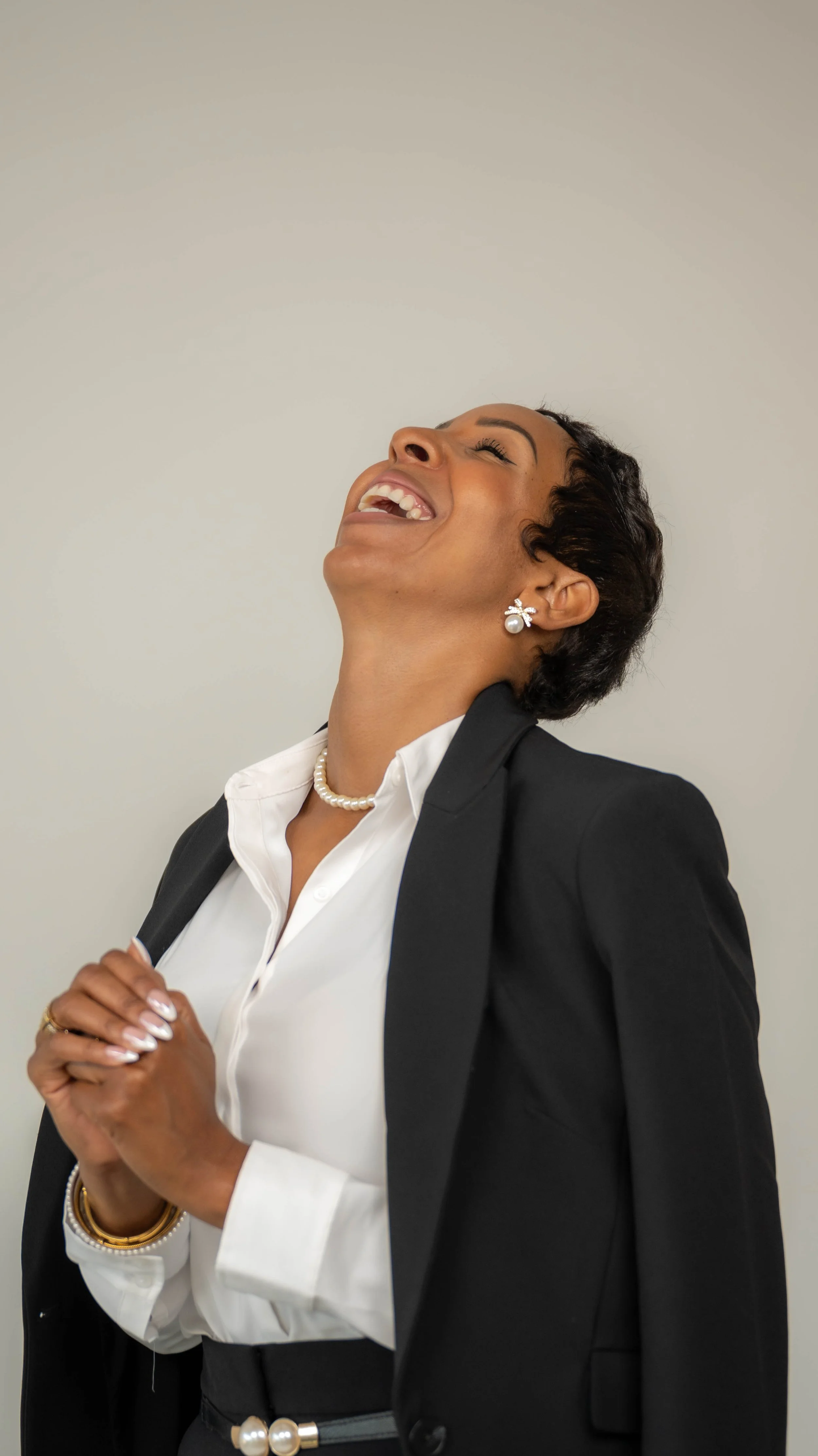 A woman with short dark hair is wearing a black blazer with a white shirt, pearl jewelry, and earrings, smiling and looking upward with her hands clasped.