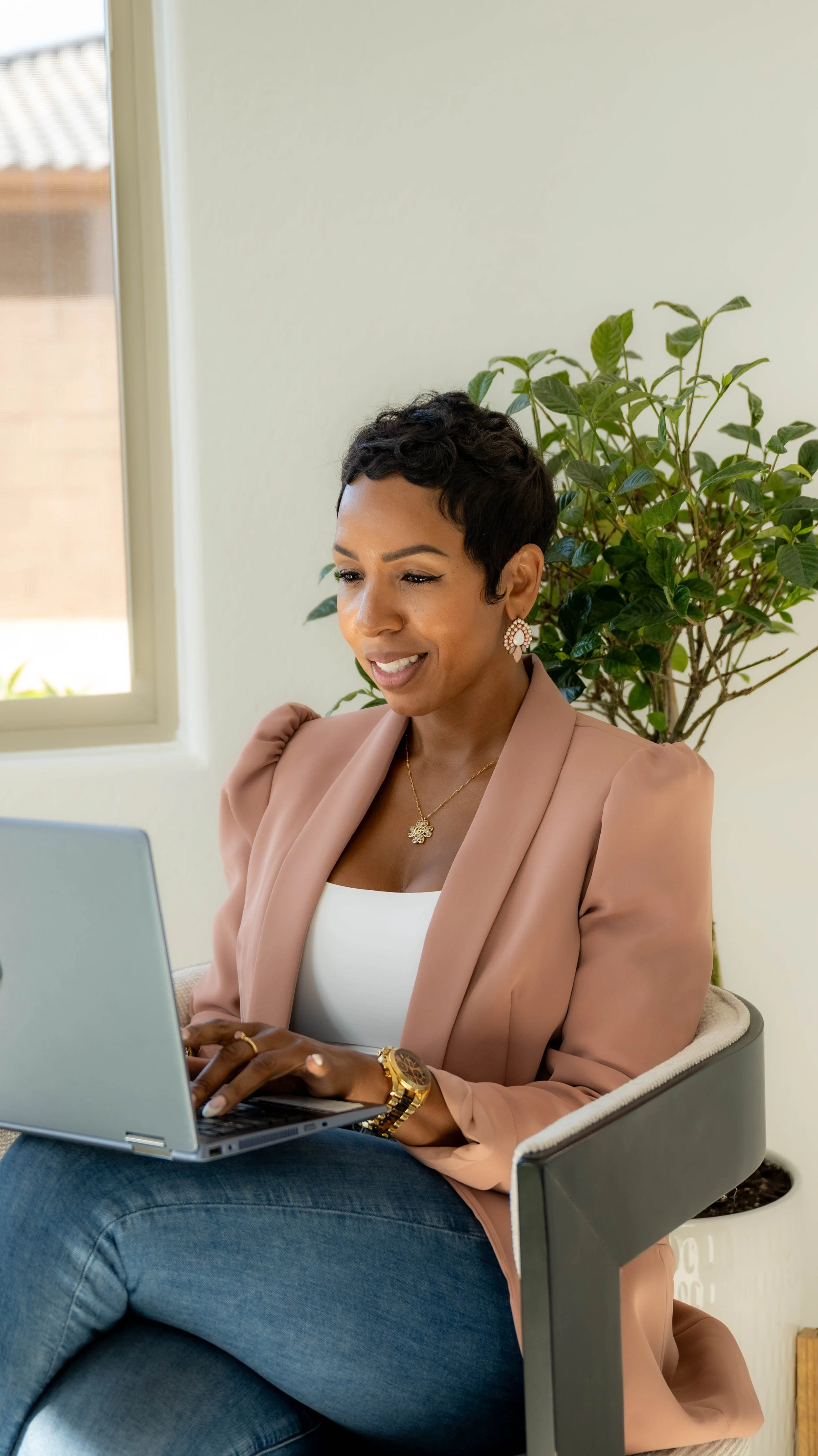 A woman with short, dark, curly hair sitting on a chair, working on a laptop, indoors near a window with a plant behind her, smiling and dressed in a beige blazer and white top.