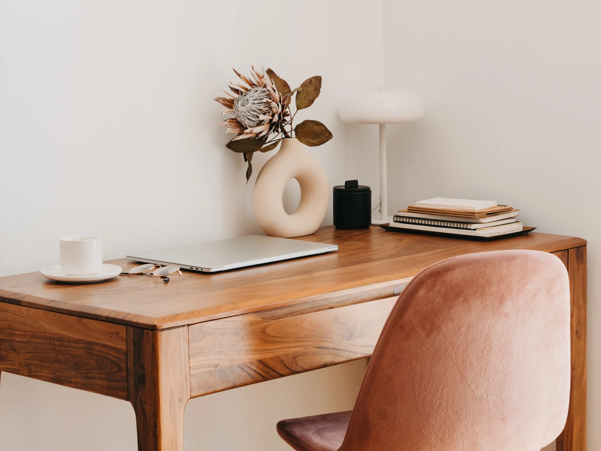 Wooden desk with a closed laptop, a white coffee mug on a saucer, a pair of glasses, a black container, a lamp, a stack of notebooks and papers, and a decorative vase with a large dried flower, against a plain white wall. Pink upholstered chair partially visible.