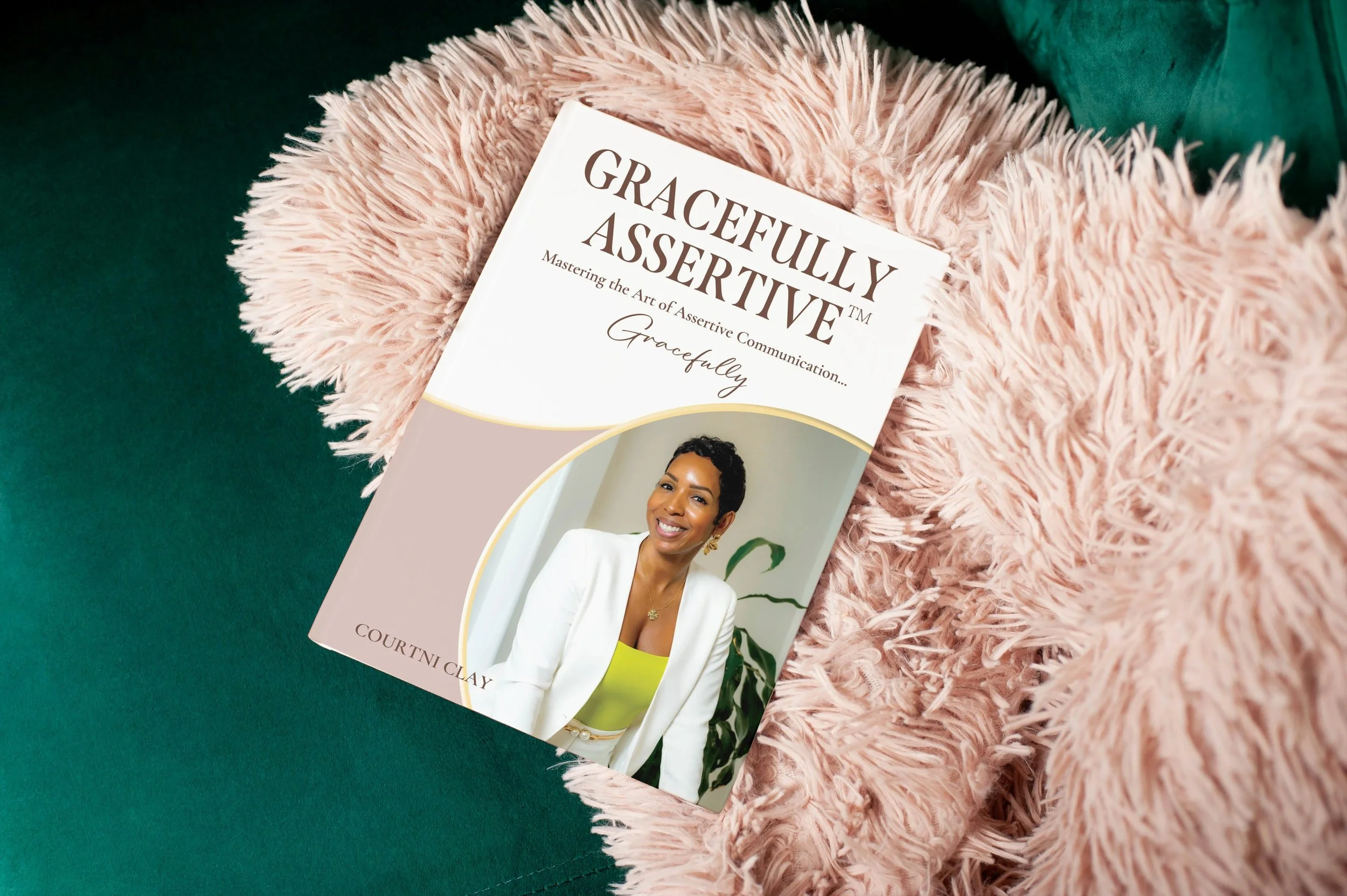 A book titled 'Gracefully Assertive: Mastering the Art of Assertive Communication' resting on a pink fluffy blanket with green fabric nearby.