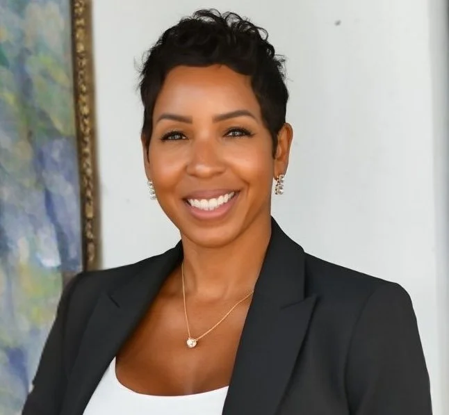 A woman with short black hair, wearing a black blazer and white top, smiling, standing indoors with a painting visible in the background.