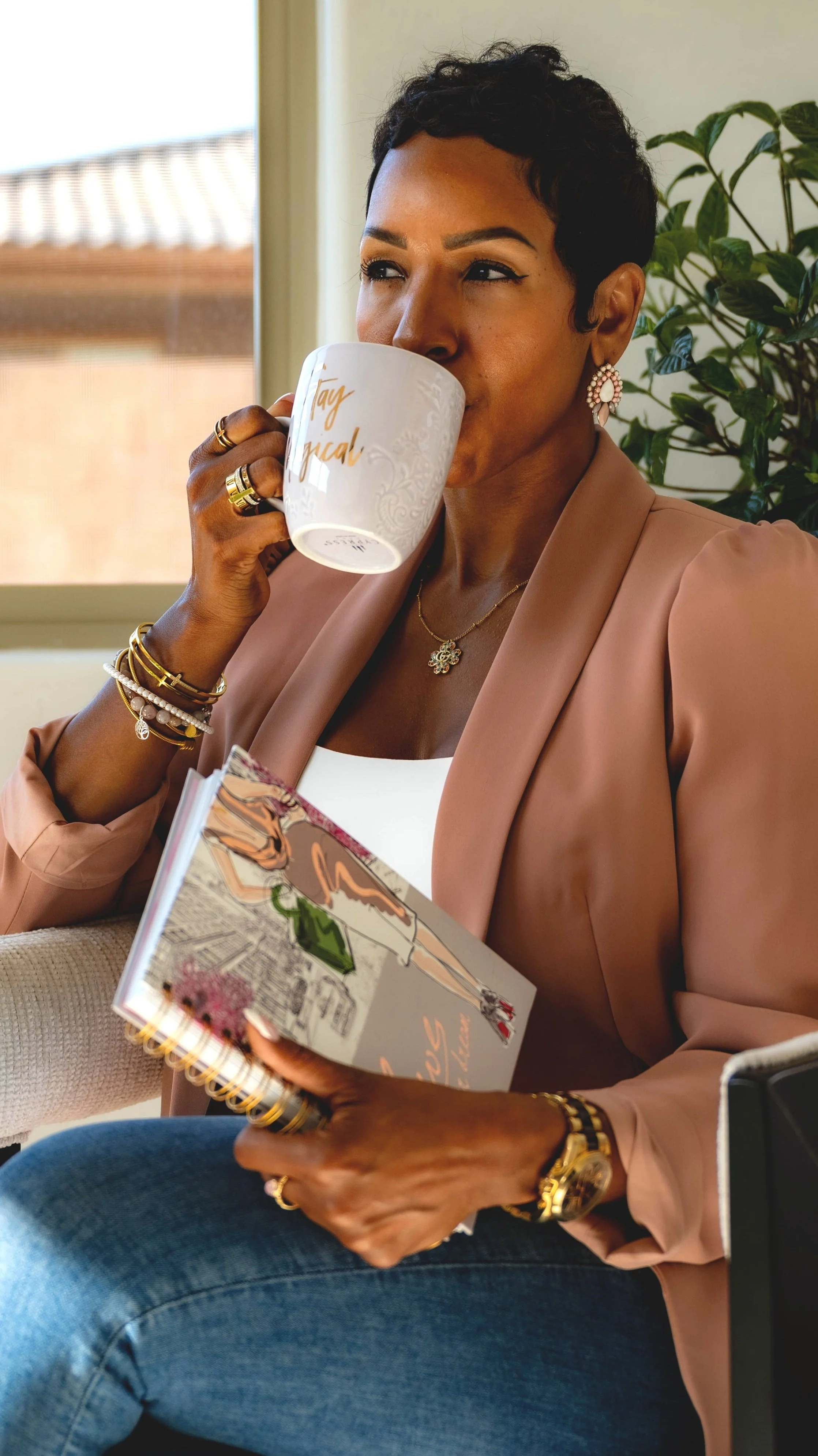 A woman with short curly black hair and gold jewelry is drinking from a white mug while holding a colorful sketchbook with fashion illustrations. She is wearing a light pink blazer, a white top, and sitting near a window with green plants in the background.
