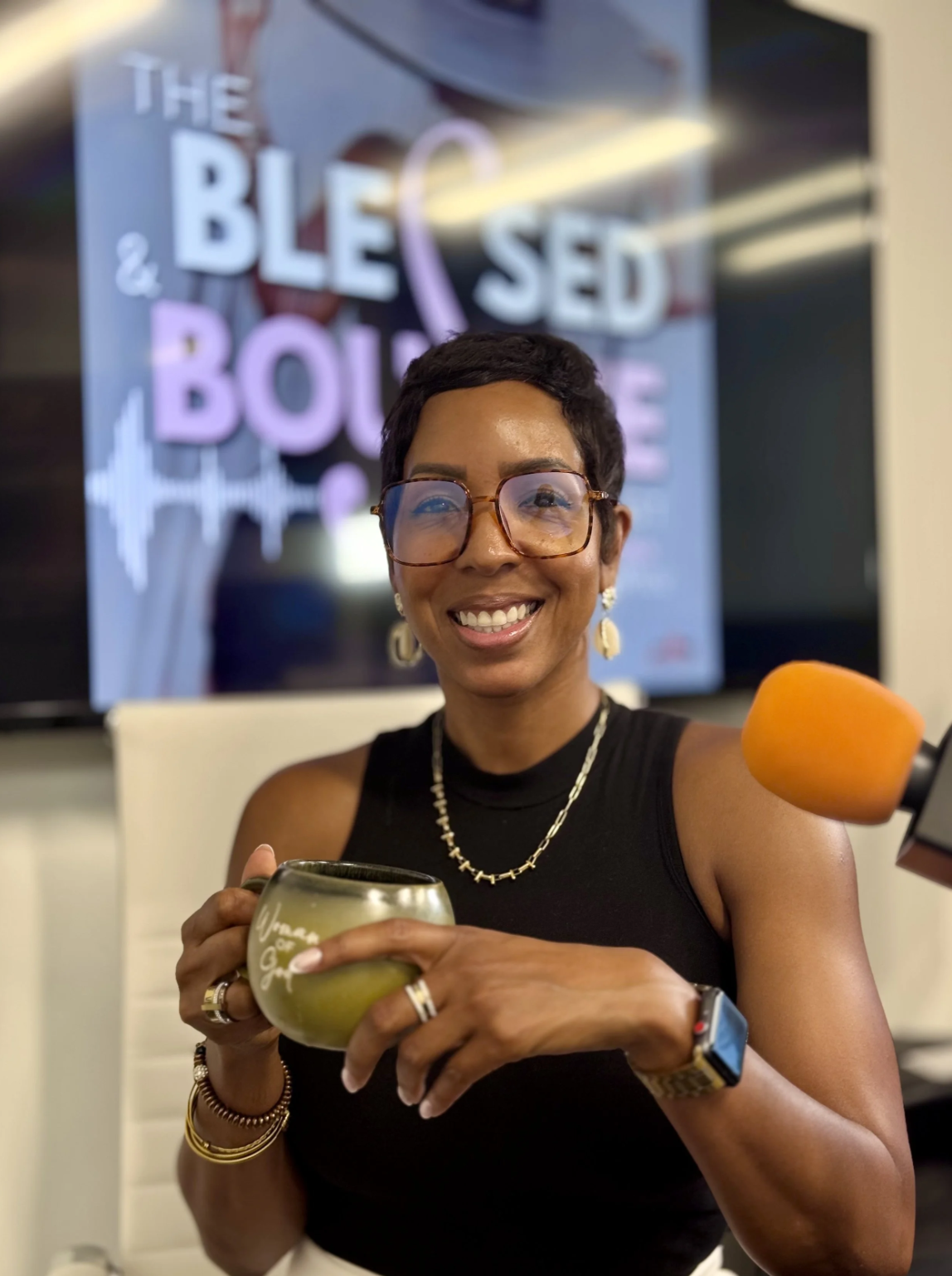 Smiling woman with short dark hair and glasses, holding a mug, during a broadcast with a microphone in foreground, and a television screen behind displaying the words 'The Blessing & The Bourse'.