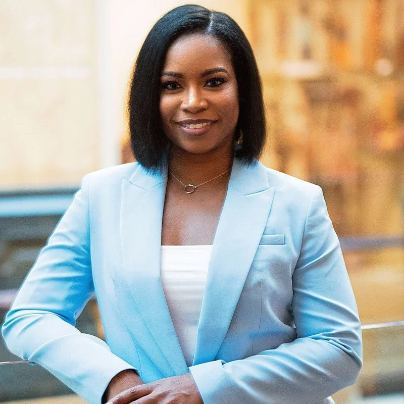 A woman with straight black hair, smiling, dressed in a light blue blazer and white top, standing indoors in front of a blurred, warmly-lit background.