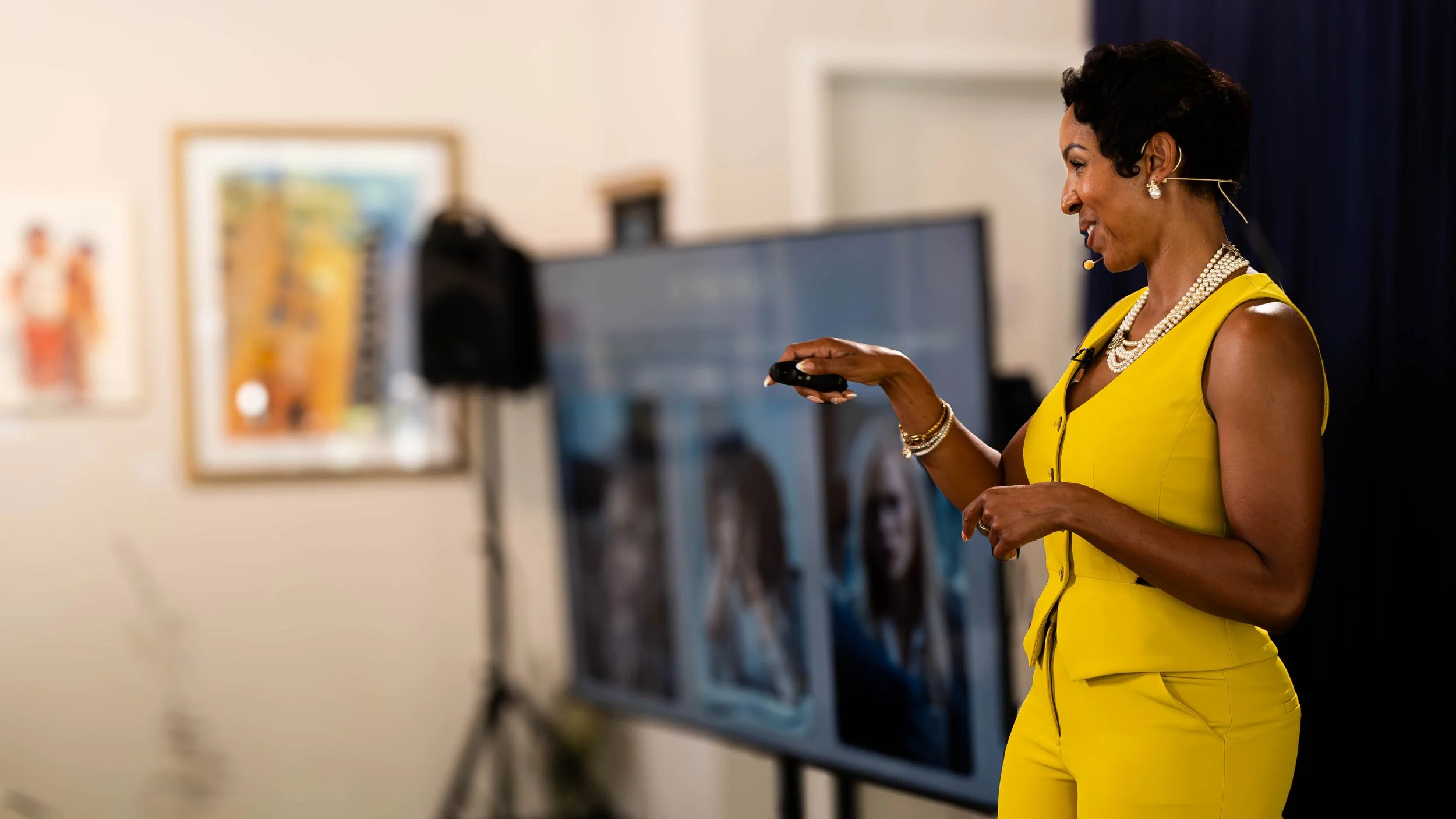 A woman in a yellow dress and pearl jewelry presenting or speaking at an event, with a blurred background of artwork and a monitor.