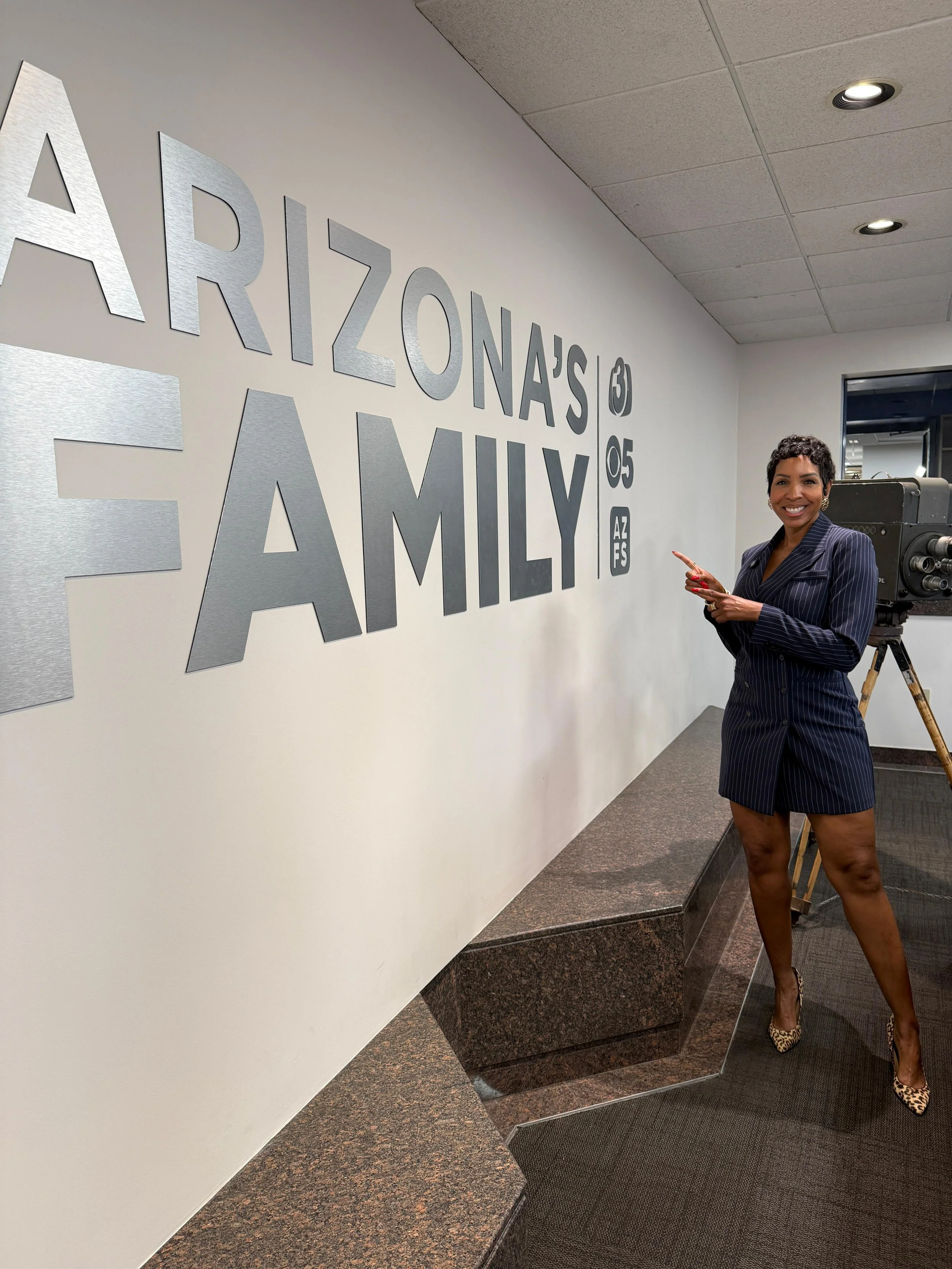 A woman in a pinstripe blazer dress and leopard print high heels standing at the Arizona's Family station, pointing towards the sign on the wall.