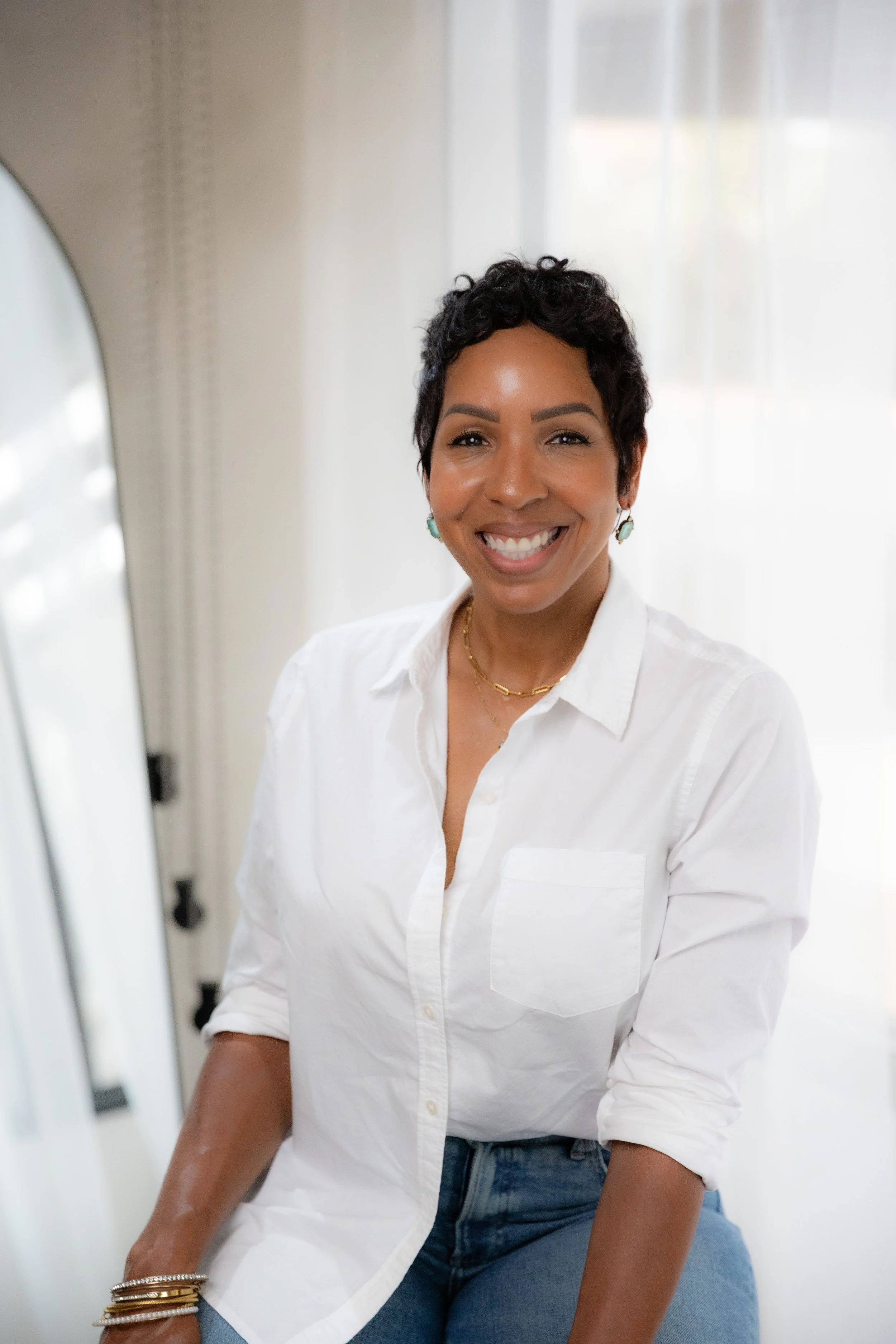 A woman smiling, wearing a white shirt and blue jeans, sitting near a mirror with a bright background.