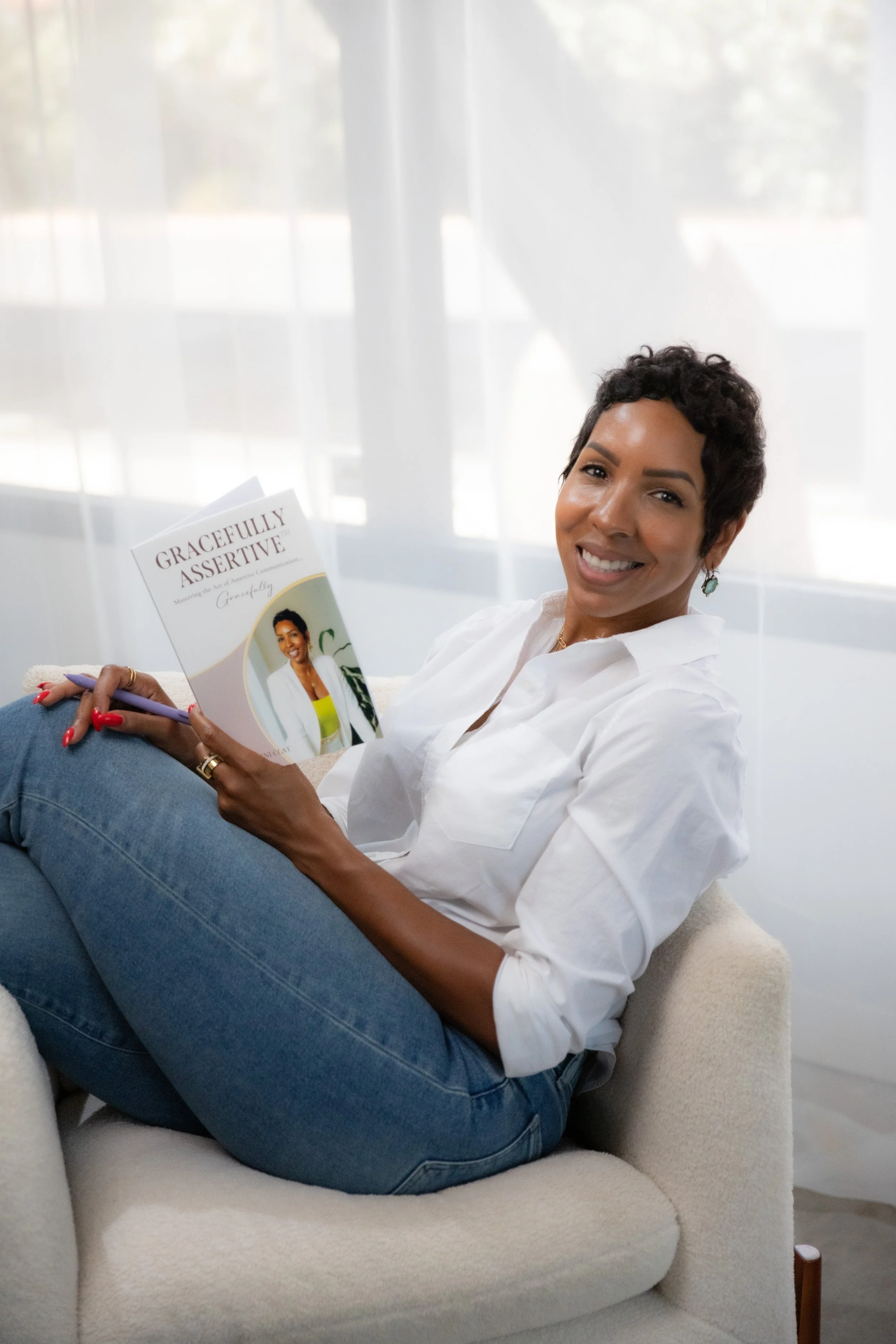 A woman with short dark hair, smiling, and wearing a white shirt and blue jeans, sitting on a beige armchair. She is holding a book titled 'Gracefully Assertive' and a purple pen, with a bright background of a window with sheer curtains.