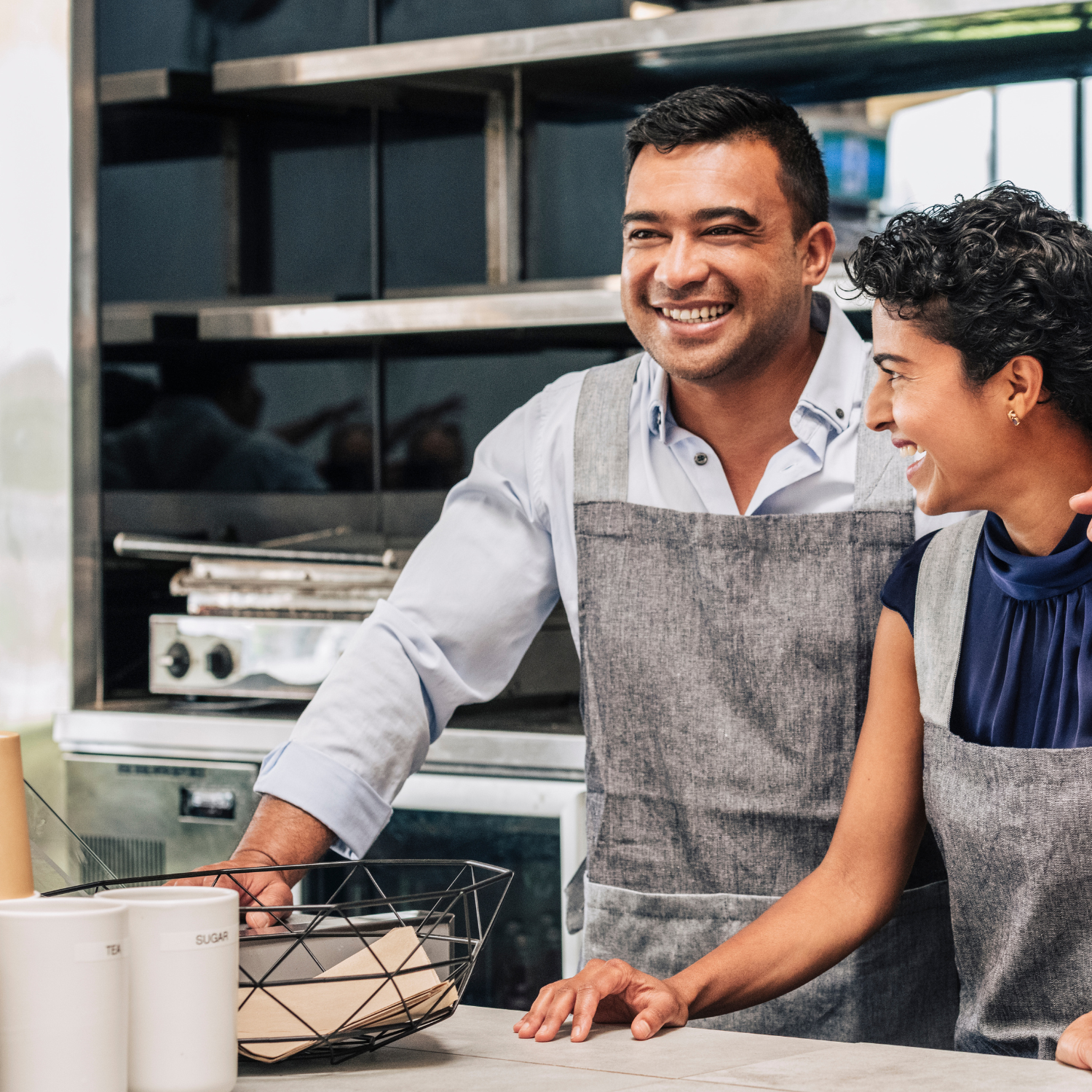 A man and woman wearing aprons, smiling in a kitchen or bakery environment.