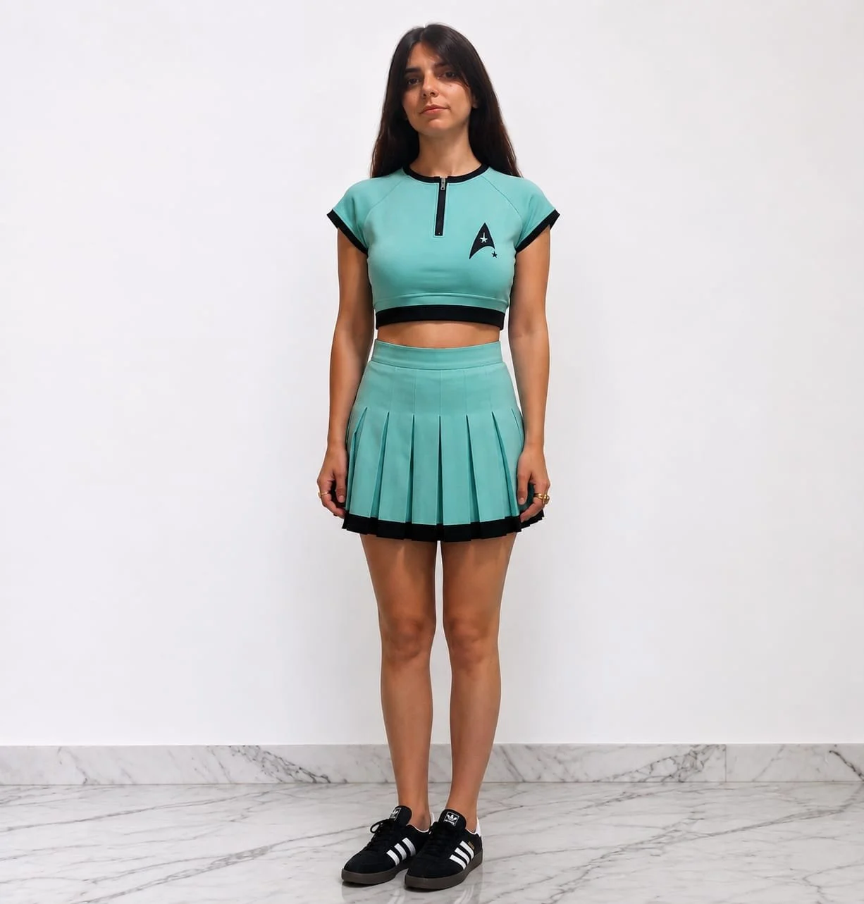 Mujer joven con peinado largo y oscuro, vestida con ropa de Star Trek, que incluye un top corto y falda plisada de color turquesa con detalles en negro, usando zapatillas deportivas negras con rayas blancas.
