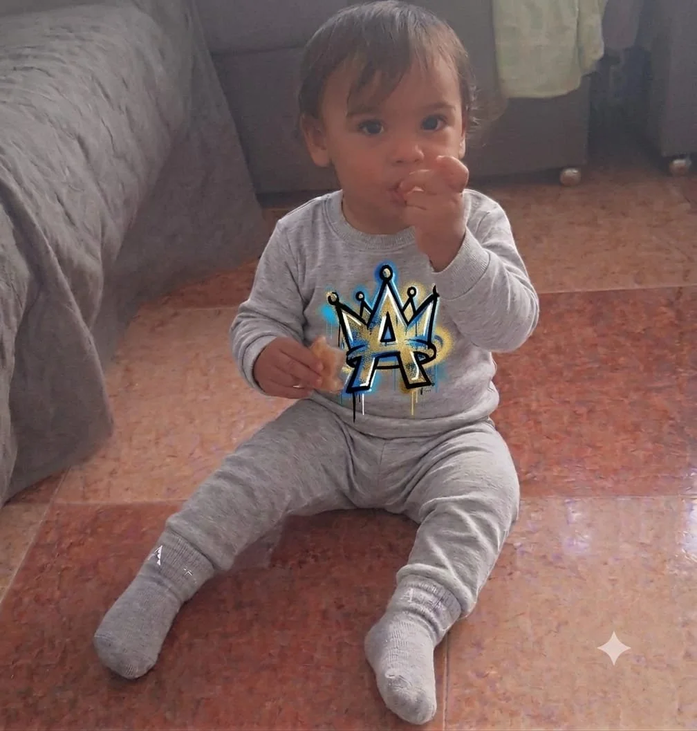 Niño pequeño sentado en el piso de baldosas, usando ropa gris, comiendo y con un dibujo de corona y letra A en su camiseta