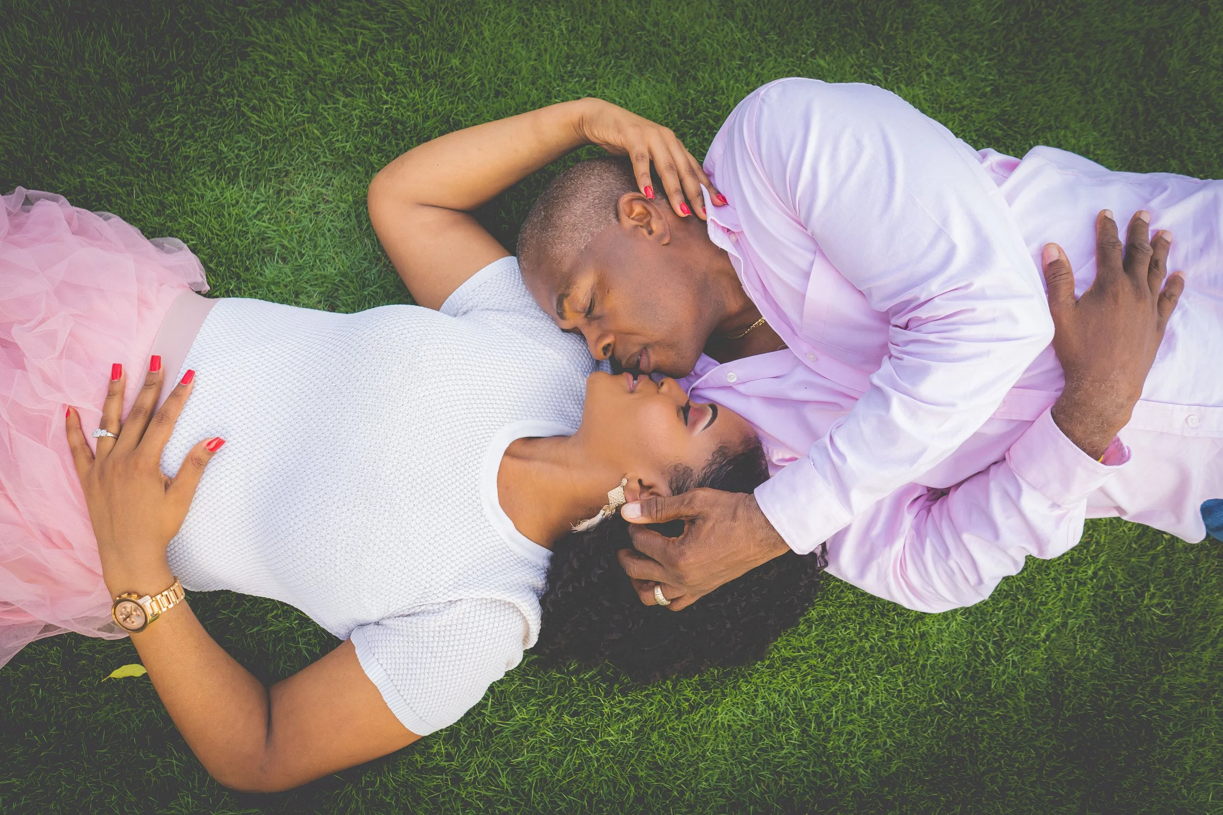 A couple is lying on the grass, nose to nose, kissing. The woman is wearing a pink skirt, a white top, and a gold watch, while the man is dressed in a light pink shirt and beige pants. The woman has red nail polish and the man has a gold chain around