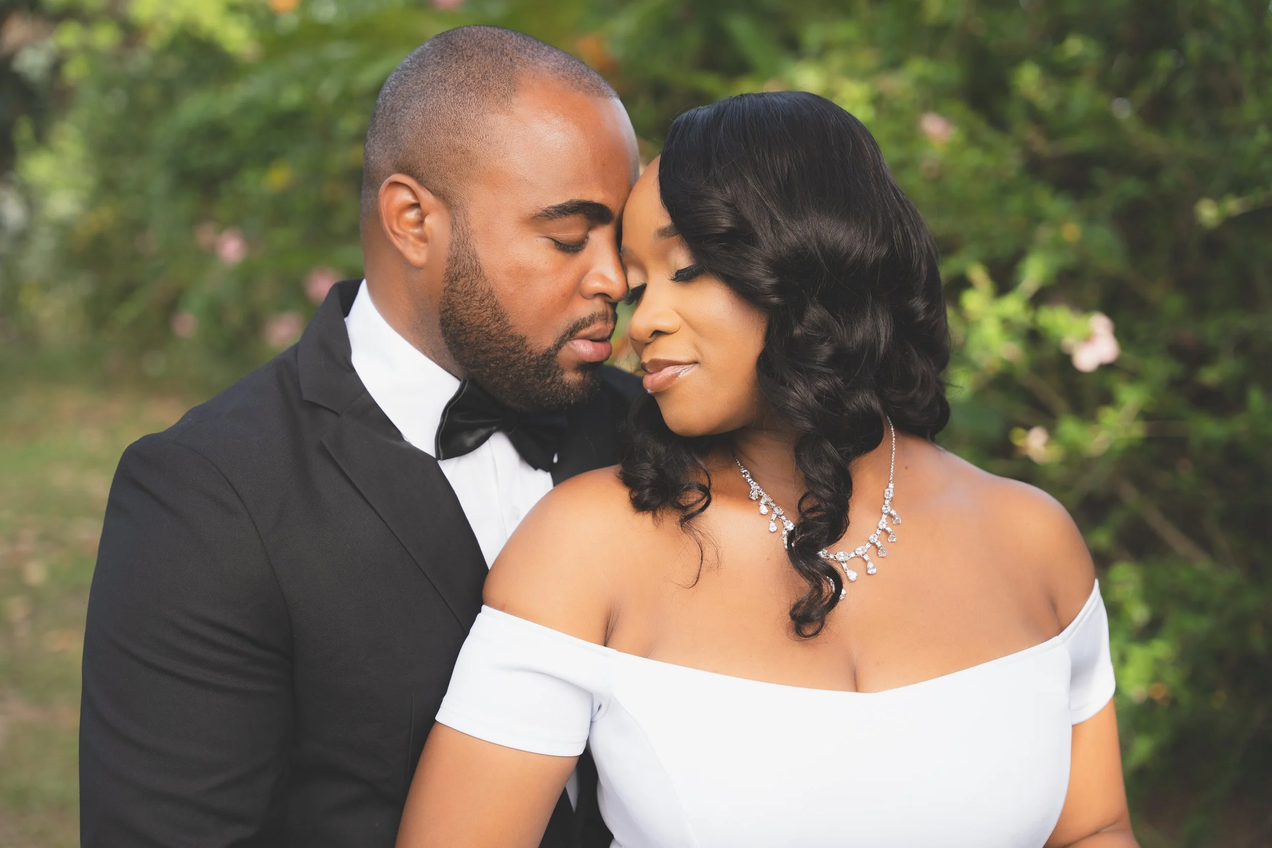 A bride and groom with eyes closed, touching foreheads in a tender moment outdoors, blurred greenery in background.