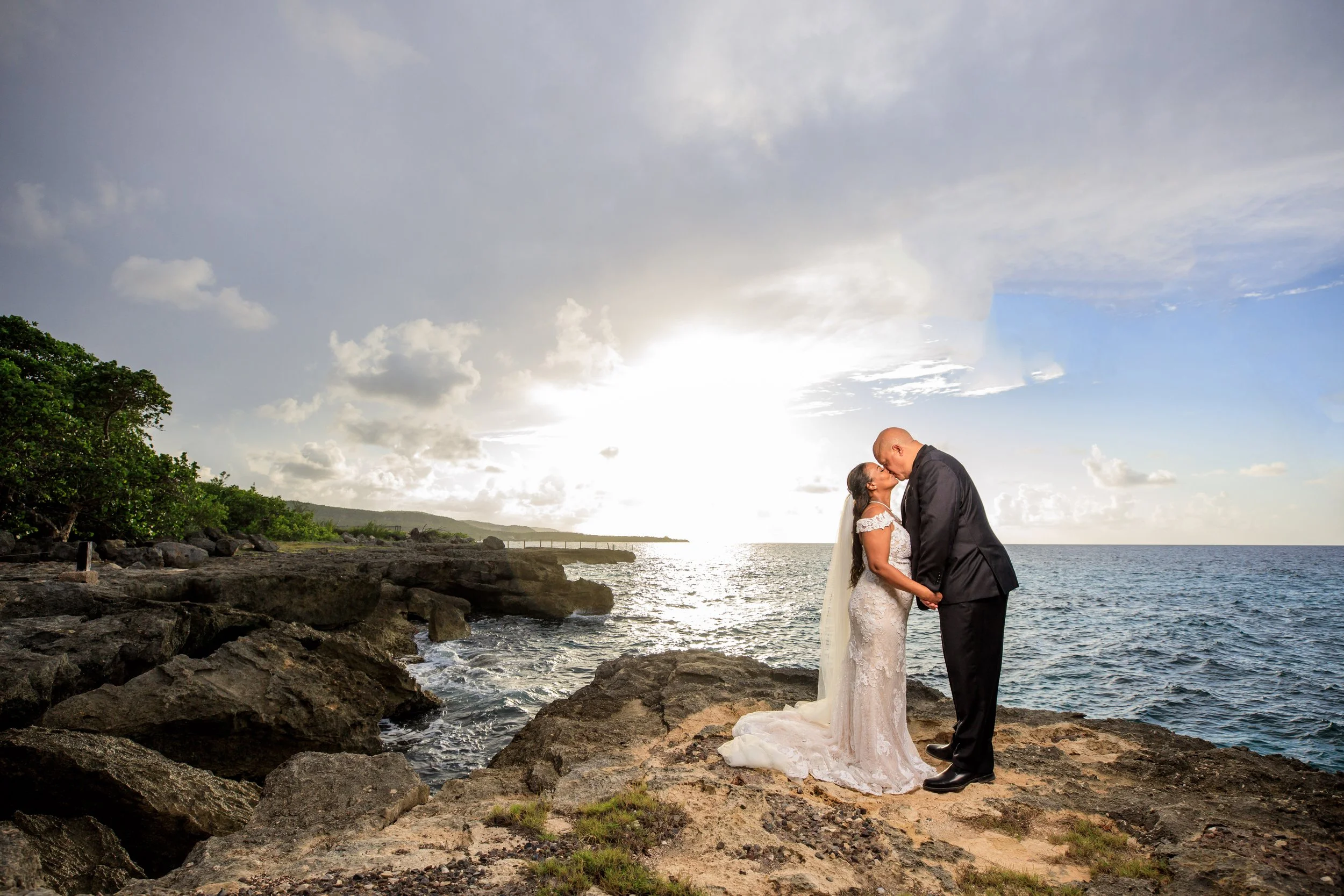 A bride and groom sharing a kiss on a rocky shoreline during sunset, with cloudy skies and the ocean in the background.