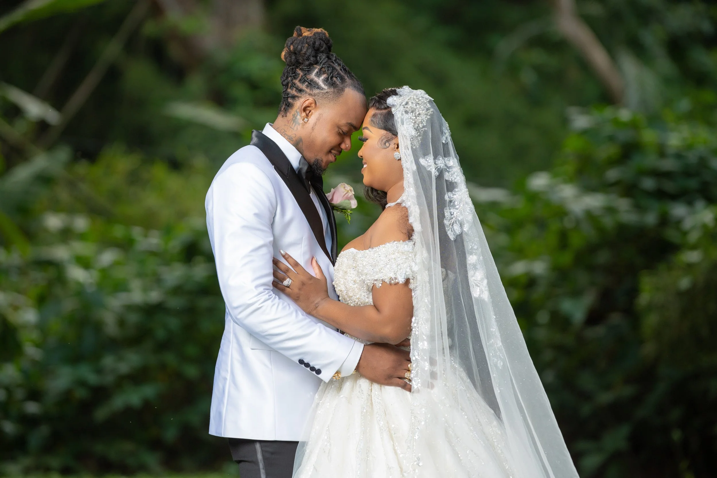 A bride and groom embrace each other outdoors, their foreheads touching and eyes closed, surrounded by greenery.