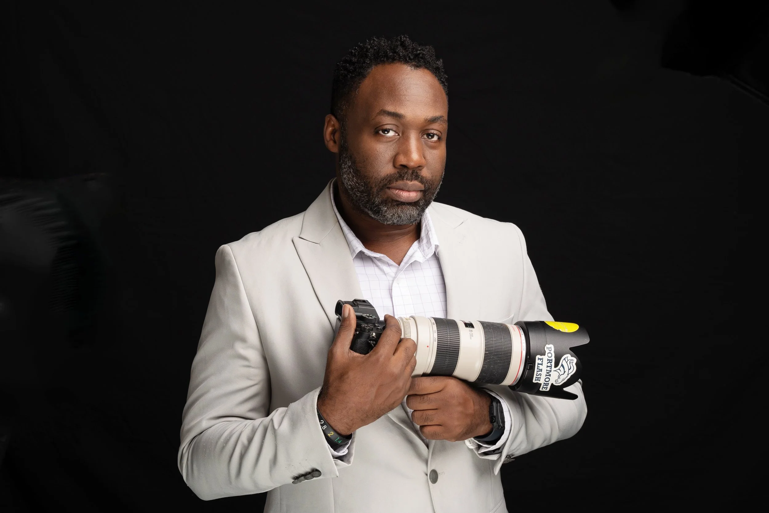 A man with dark curly hair and a beard wearing a white blazer and checkered shirt holding a professional camera with a large telephoto lens against a black background.