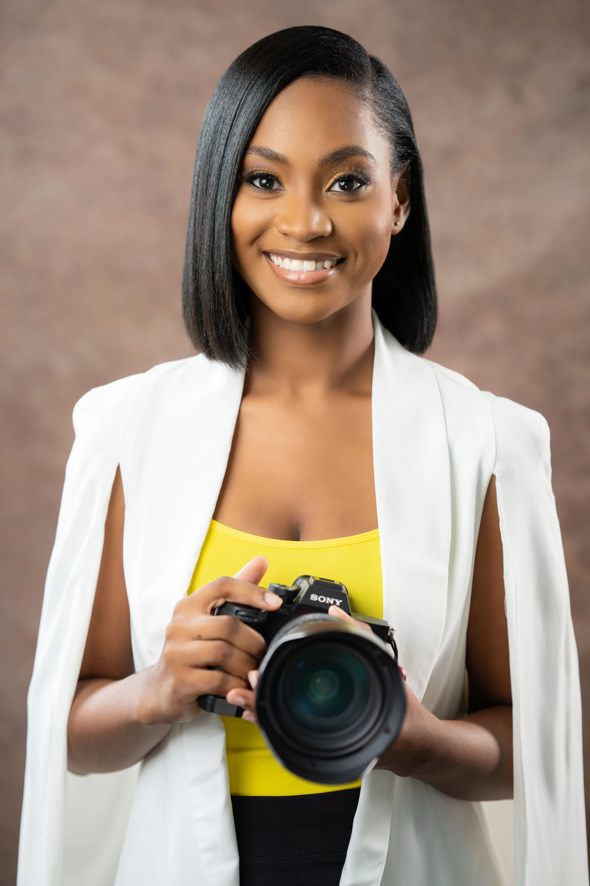A young woman with straight black hair, wearing a white blazer over a yellow top, smiling and holding a Sony camera.