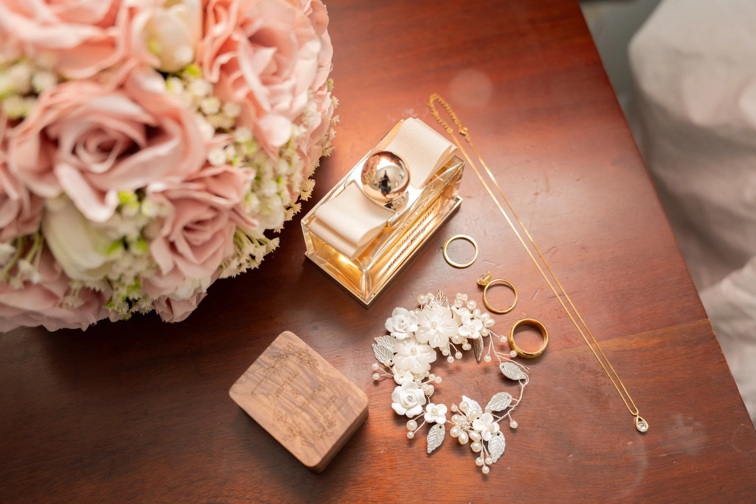 A wedding ring, jewelry, and a bouquet of pink and white flowers on a wooden surface.