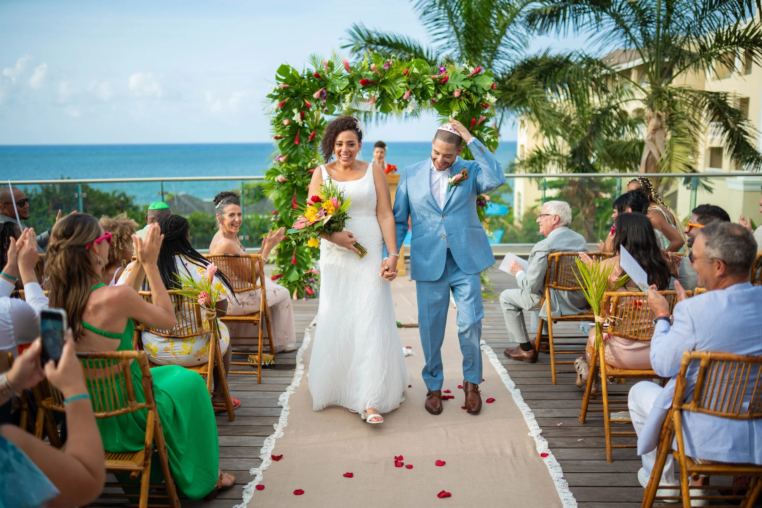 A joyful couple walking down the aisle at a wedding ceremony on a rooftop terrace with ocean view, surrounded by guests clapping and taking photos, tropical plants, and floral decorations.