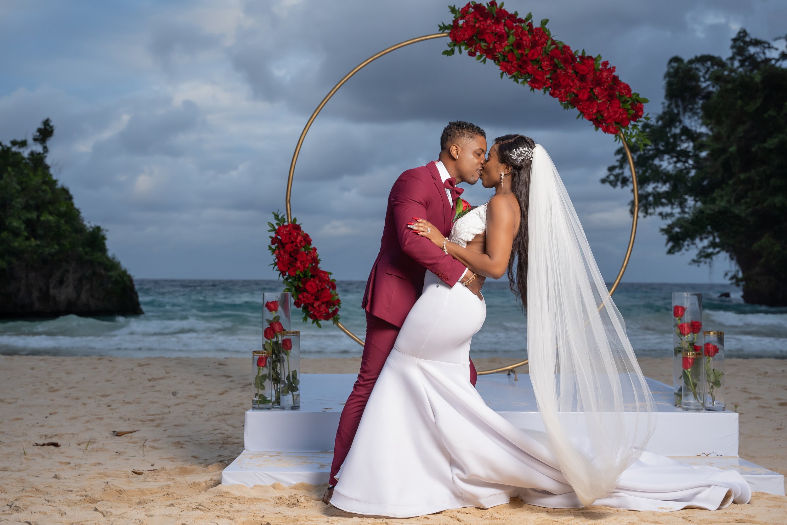 A newlywed couple sharing a kiss at a beach wedding ceremony during daytime, with a circular floral arch decorated with red flowers and greenery behind them, ocean waves in the background, and vases with red roses on a white platform.