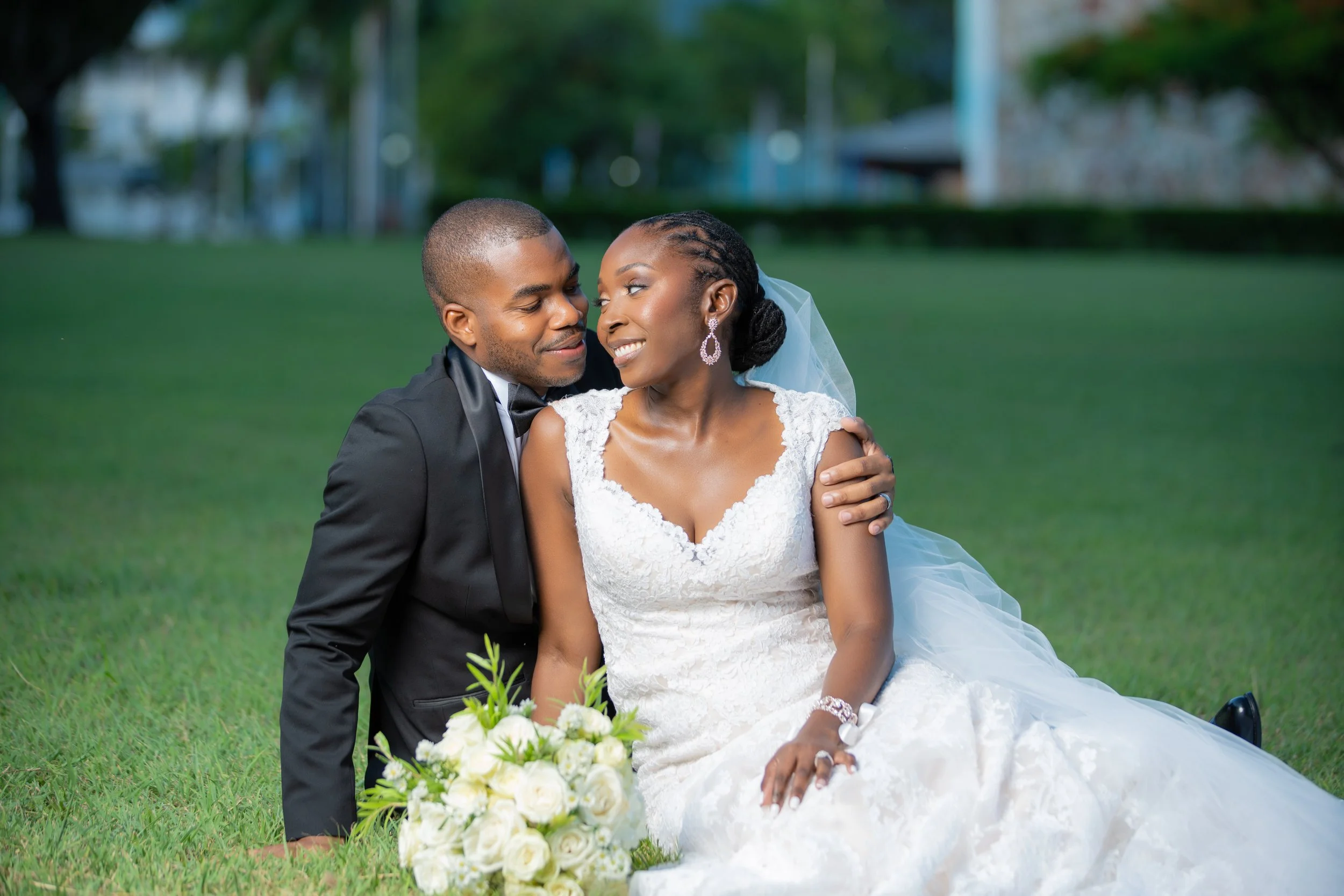 A newlywed couple sitting on grass in a park, smiling at each other, with a bouquet of white roses in front of them.