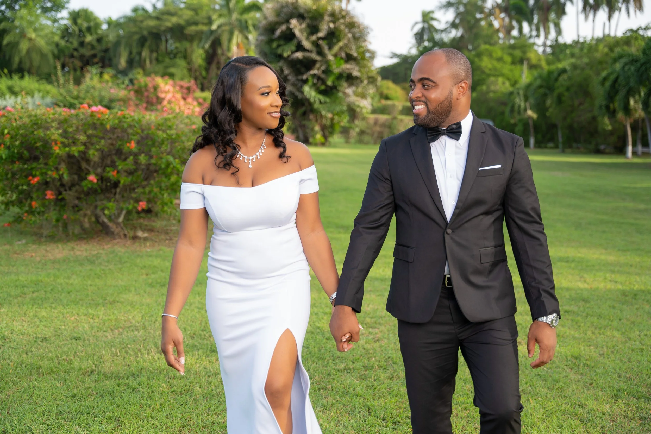 A couple dressed in formal wedding attire walking hand in hand outdoors in a lush garden setting with trees and flowering bushes, smiling at each other.