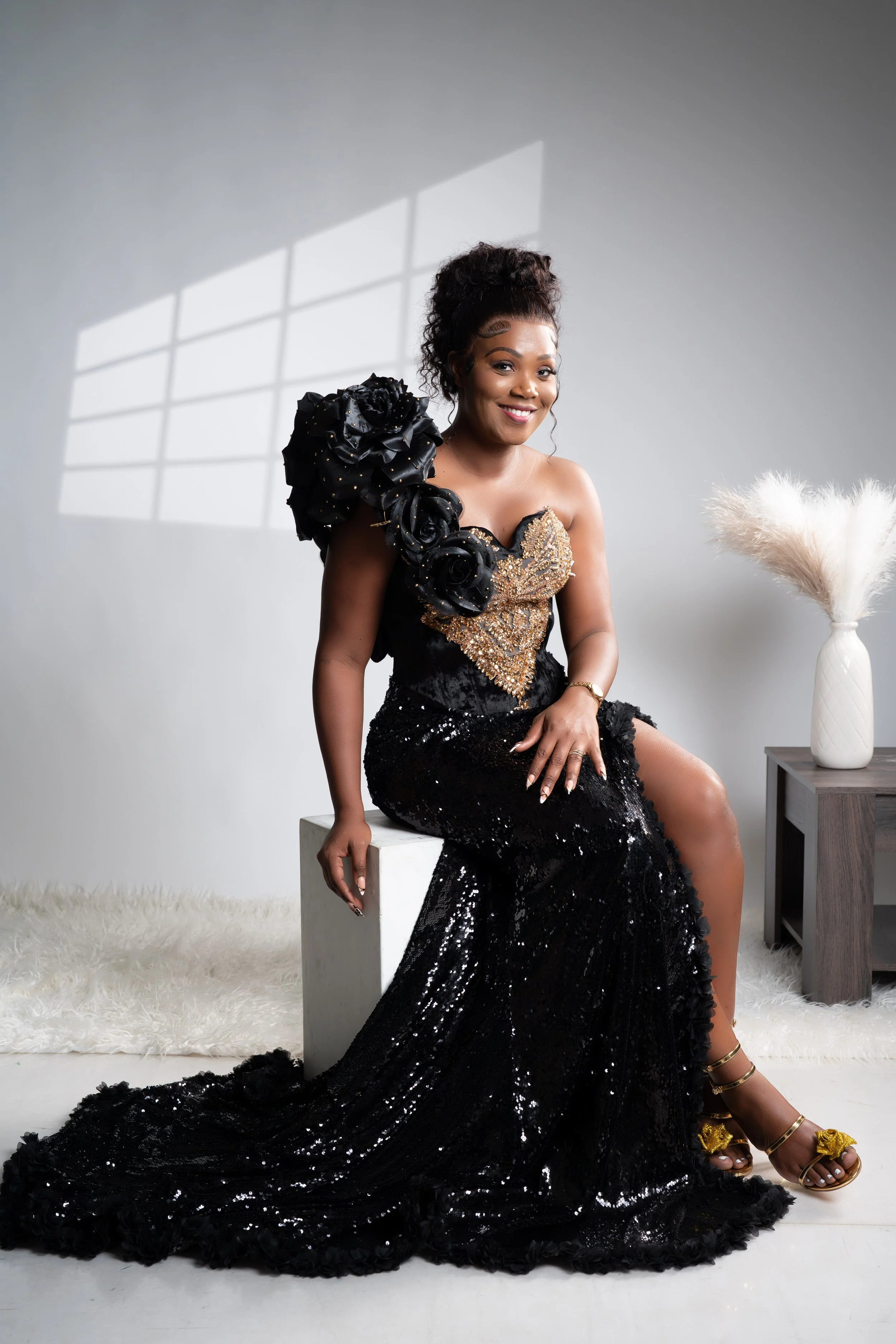 A woman in a glamorous black sequin gown with gold embellishments, sitting on a white box in a minimal indoor setting, smiling, with a white faux fur rug, a white vase with pampas grass, and a window casting light on the wall.