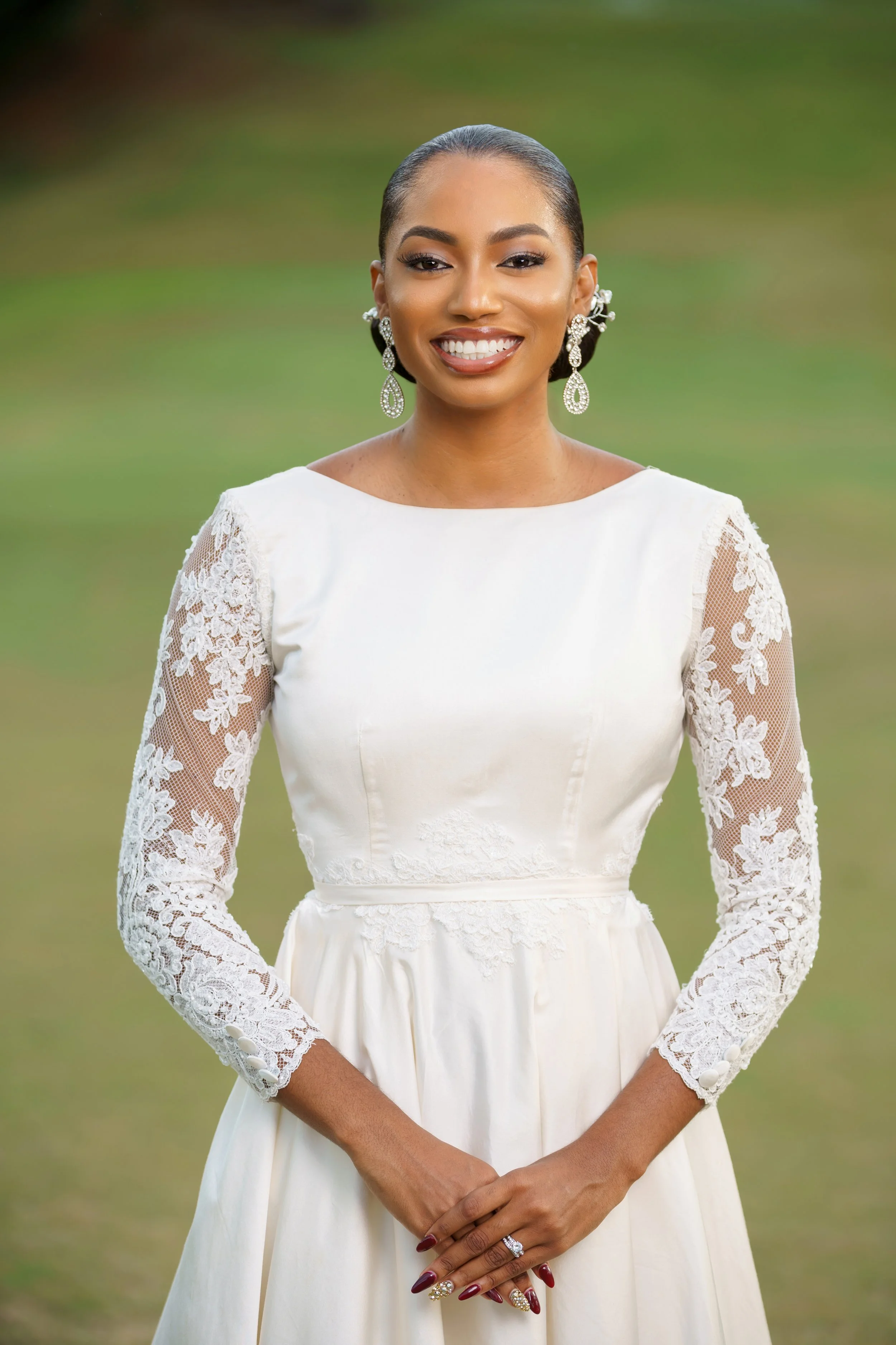 A woman in a white wedding dress with lace sleeves and floral embroidery, smiling with her hands clasped in front of her outdoors with a green background.