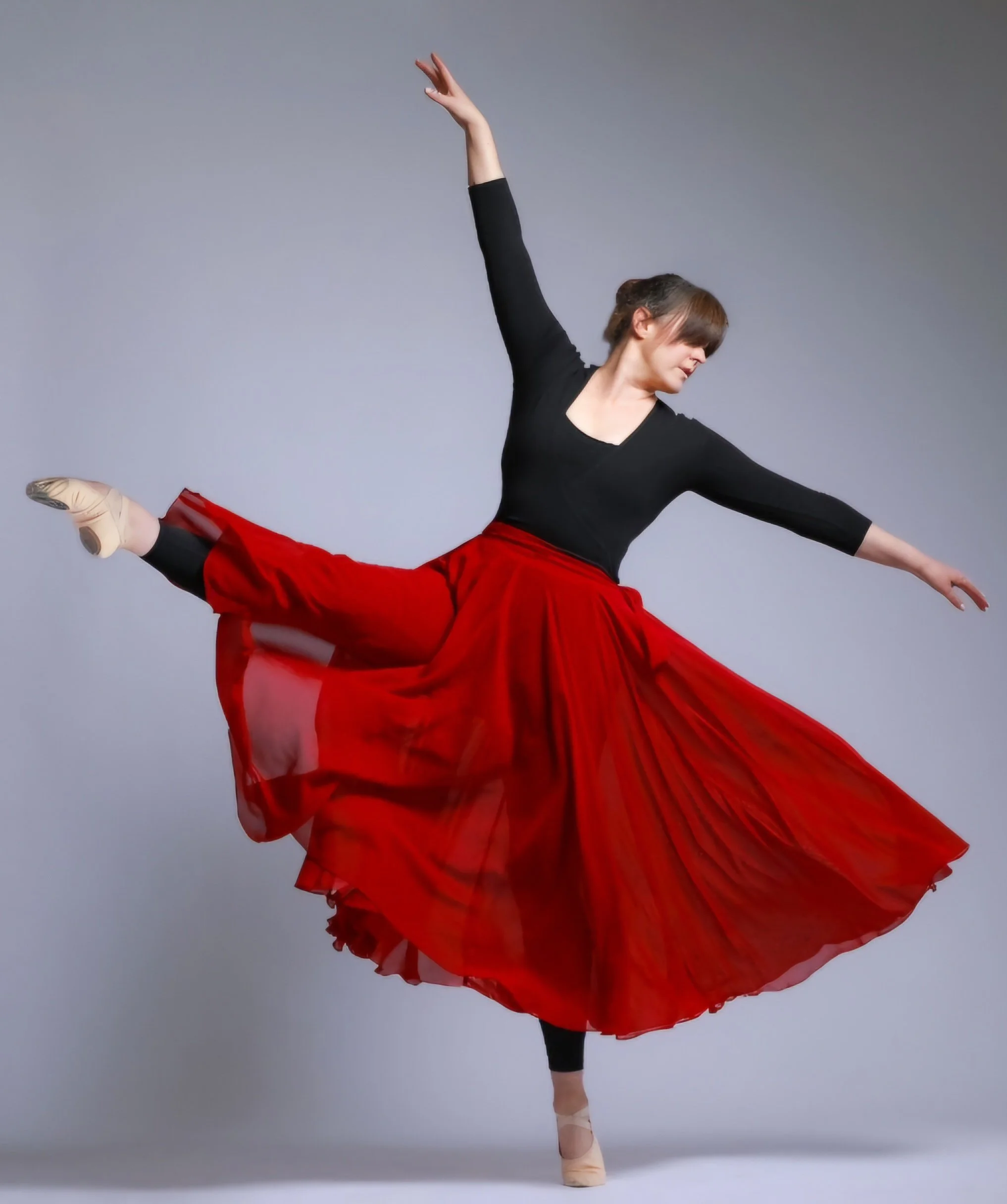 Ballet dancer in a black top and red tutu performing a ballet pose, with one leg raised behind her and arms extended, against a gray background.