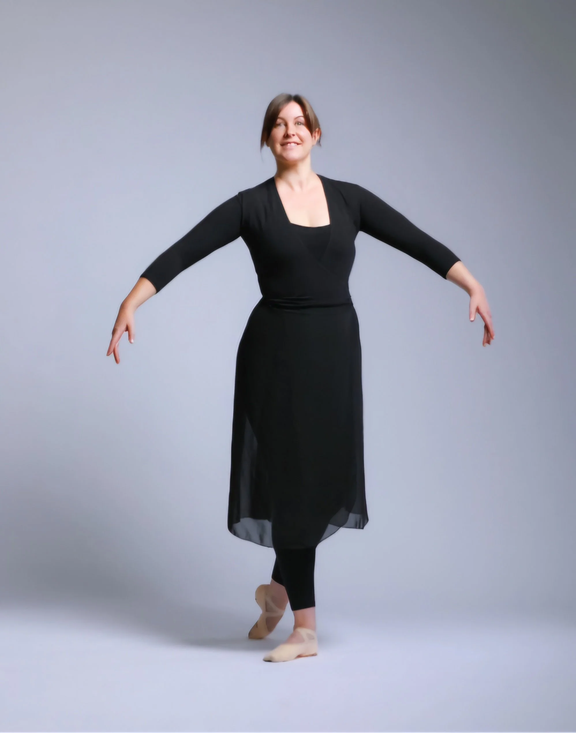 A woman ballet dancer standing en pointe on one foot, with her arms outstretched and a smile on her face, wearing a black dress and ballet shoes, against a plain light grey background.