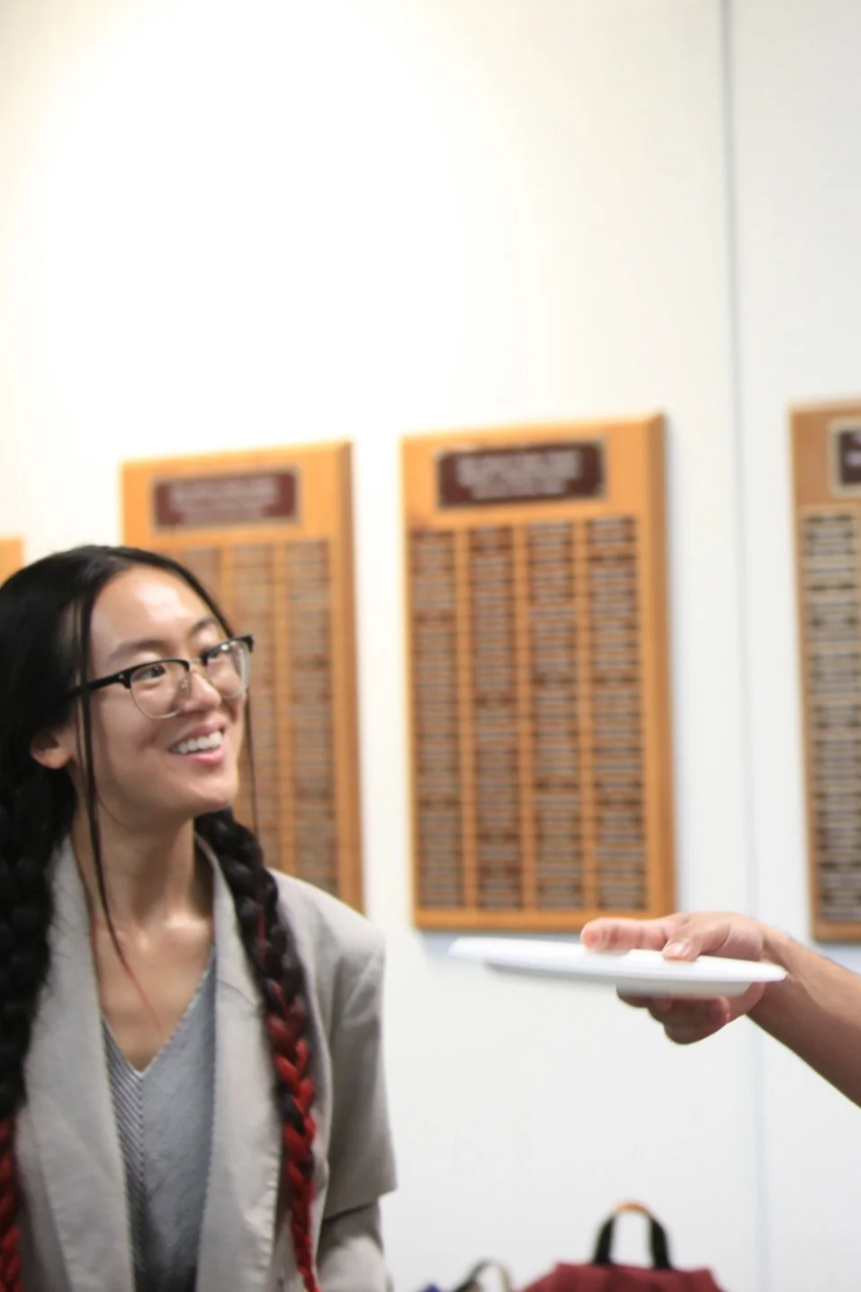 A smiling young woman with glasses and braided hair looking at a person holding a plate in a room with wooden plaques on the wall.