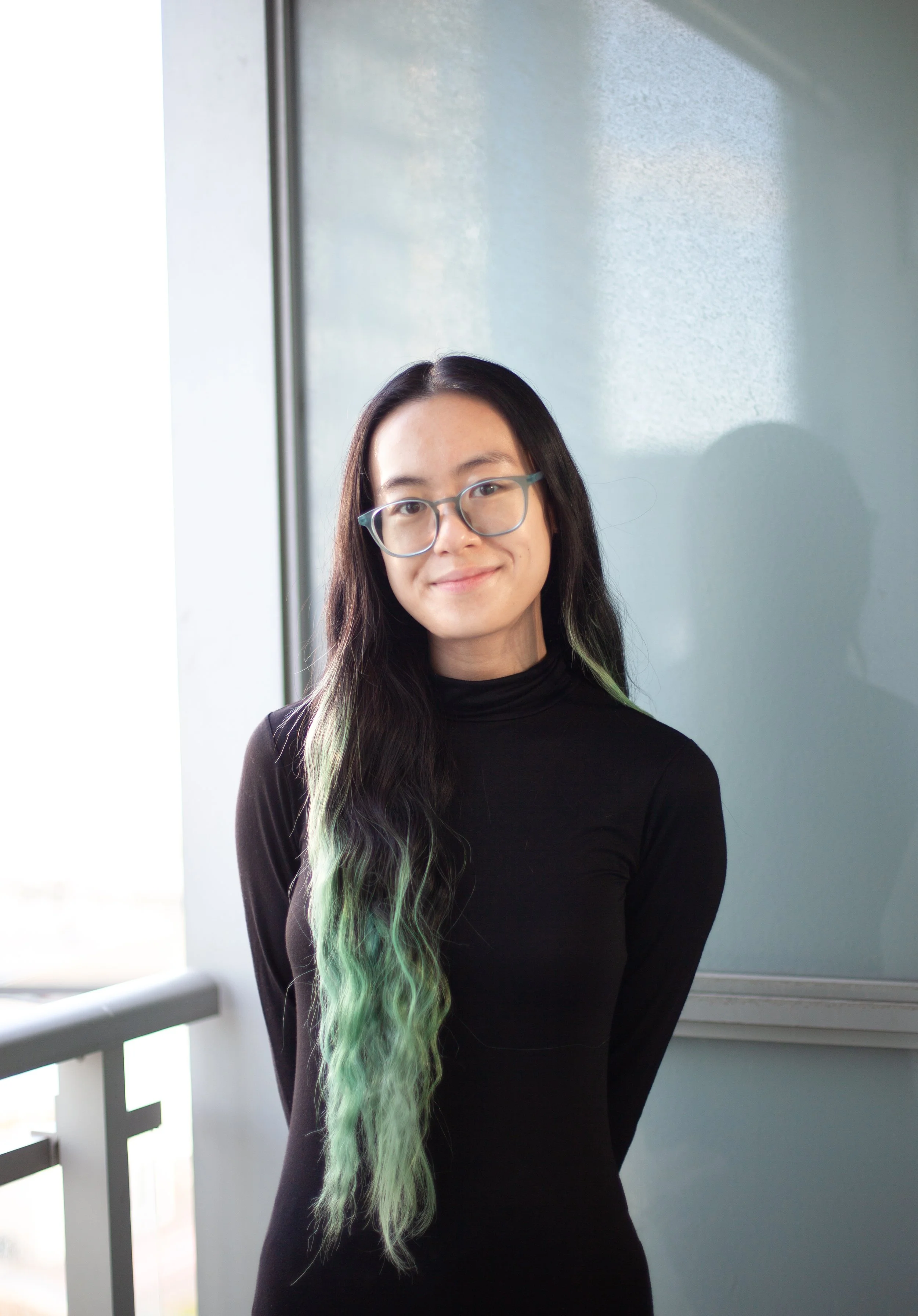 A woman with long black hair and green highlights, wearing glasses and a black turtleneck, standing near a window with natural light, smiling slightly.
