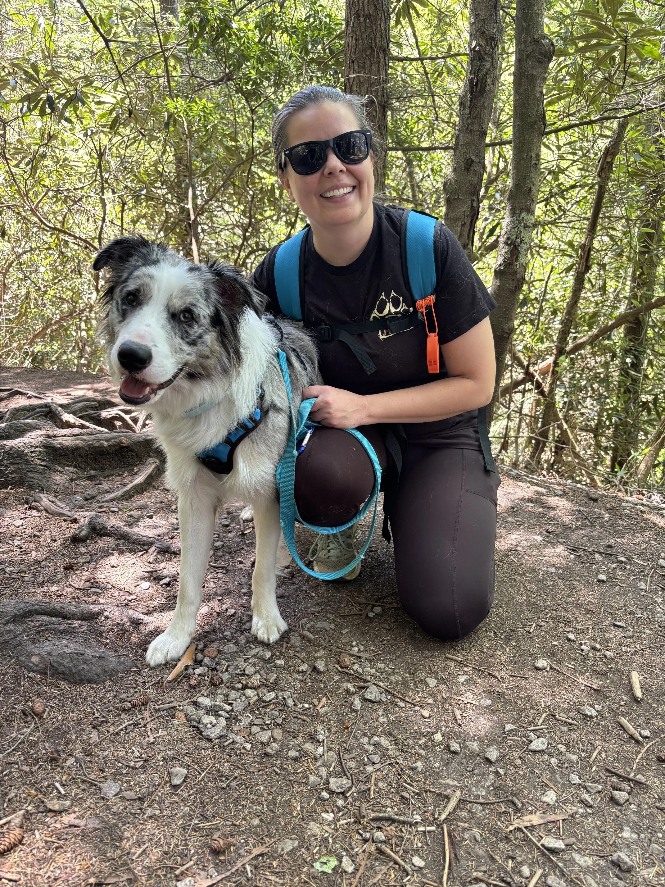 A woman kneeling on a forest trail with a blue merle Border Collie dog, both smiling. She is wearing sunglasses, a black t-shirt, and a blue backpack, surrounded by green trees and natural forest ground.
