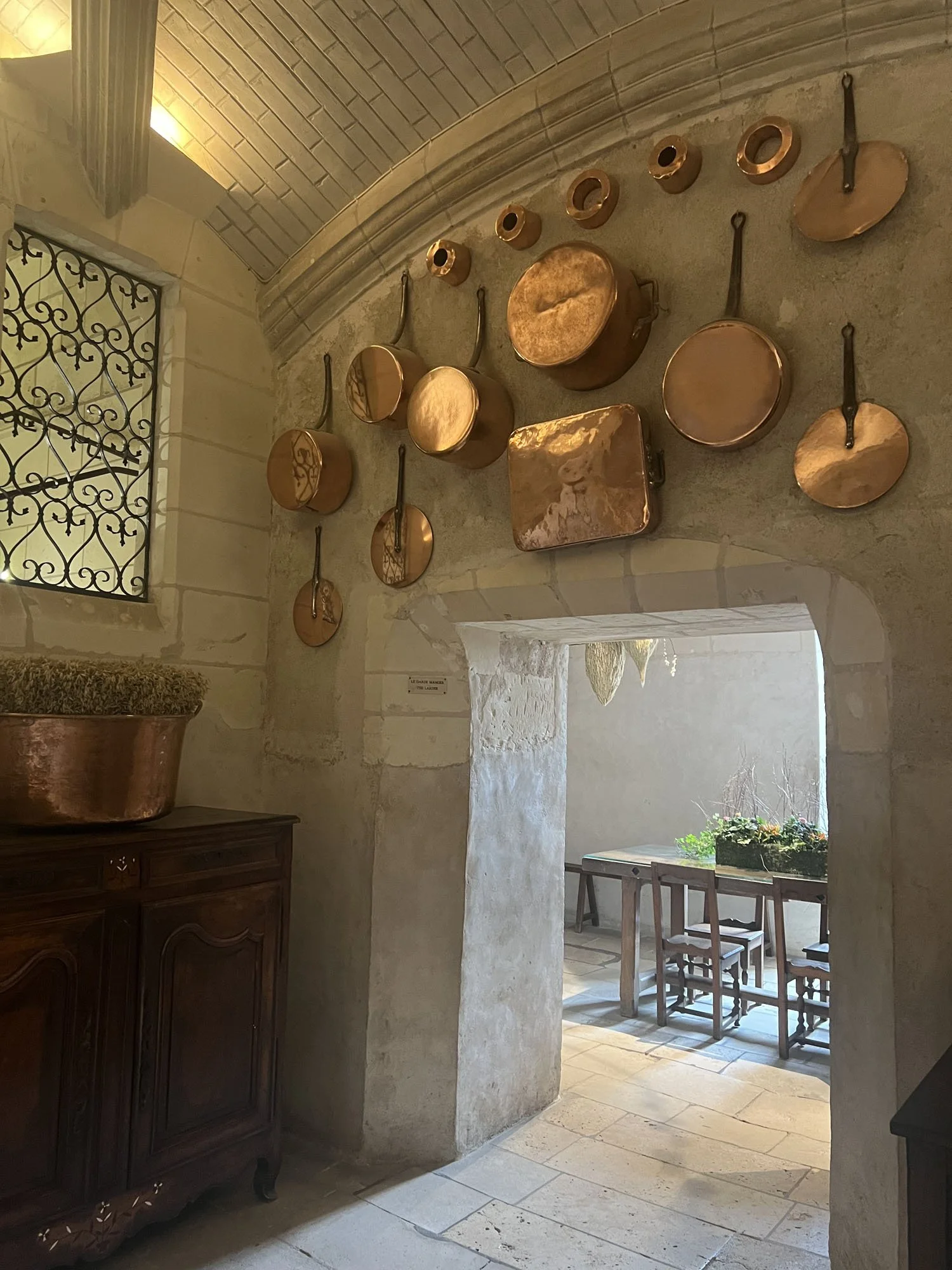 Historic stone interior with copper cookware displayed on an arched wall, antique cabinet, wrought-iron window, and dining area beyond