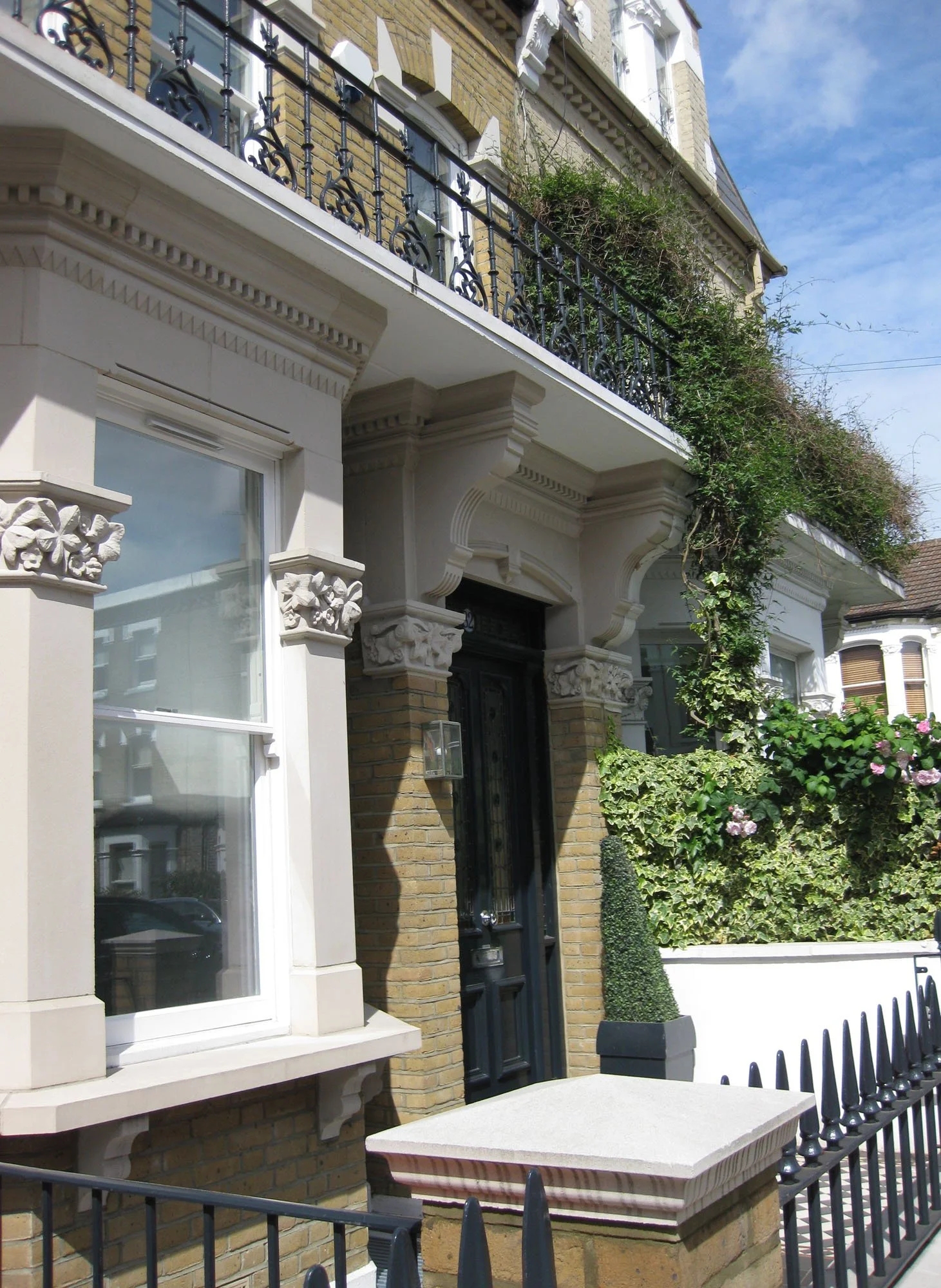 Ornate Victorian townhouse facade with brickwork, sculpted white stone details, black iron railings, balcony, and ivy-covered garden wall