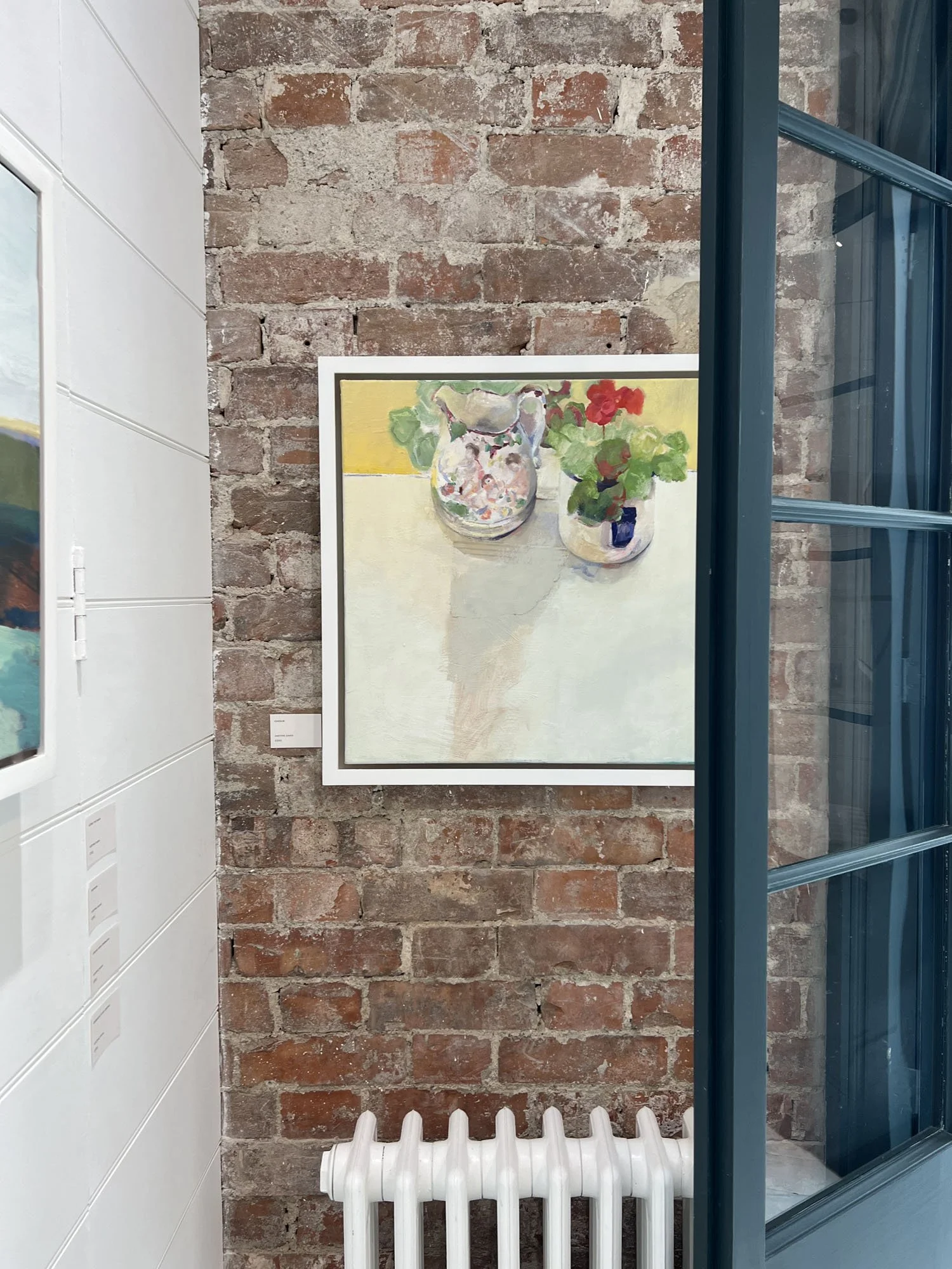 Framed still-life painting of flowers in vases displayed on an exposed brick gallery wall beside a blue-framed window and white radiator