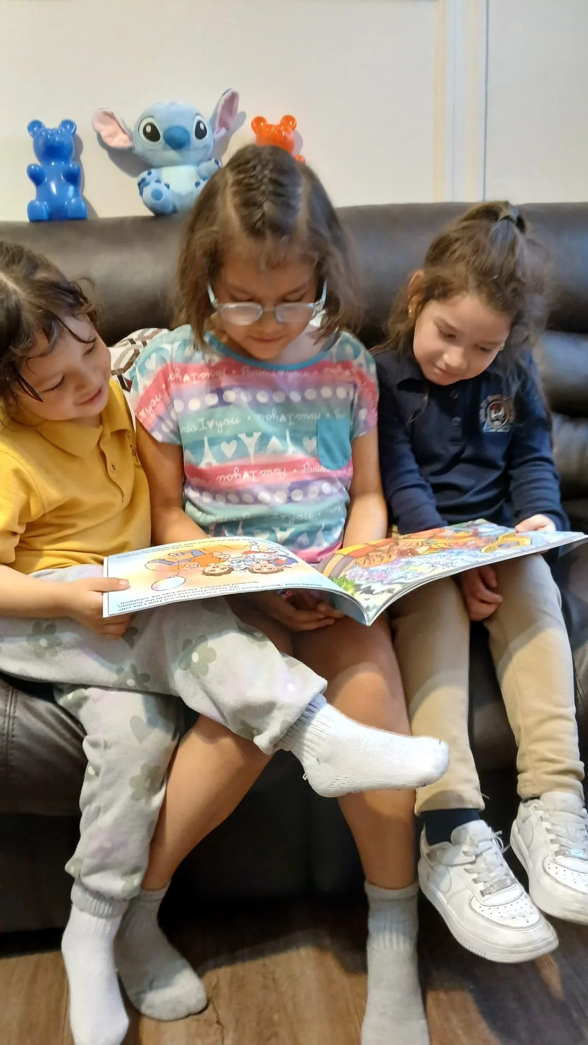 Three young girls sitting on a couch looking at a book, with a Stitch plush and two teddy bear-shaped decorations in the background.