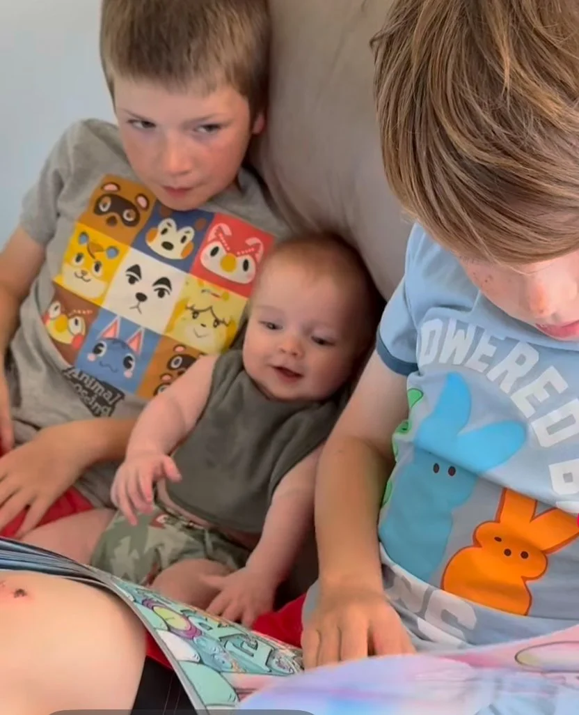 Three children, two boys and a baby, sitting on a couch and looking at a colorful book or magazine.