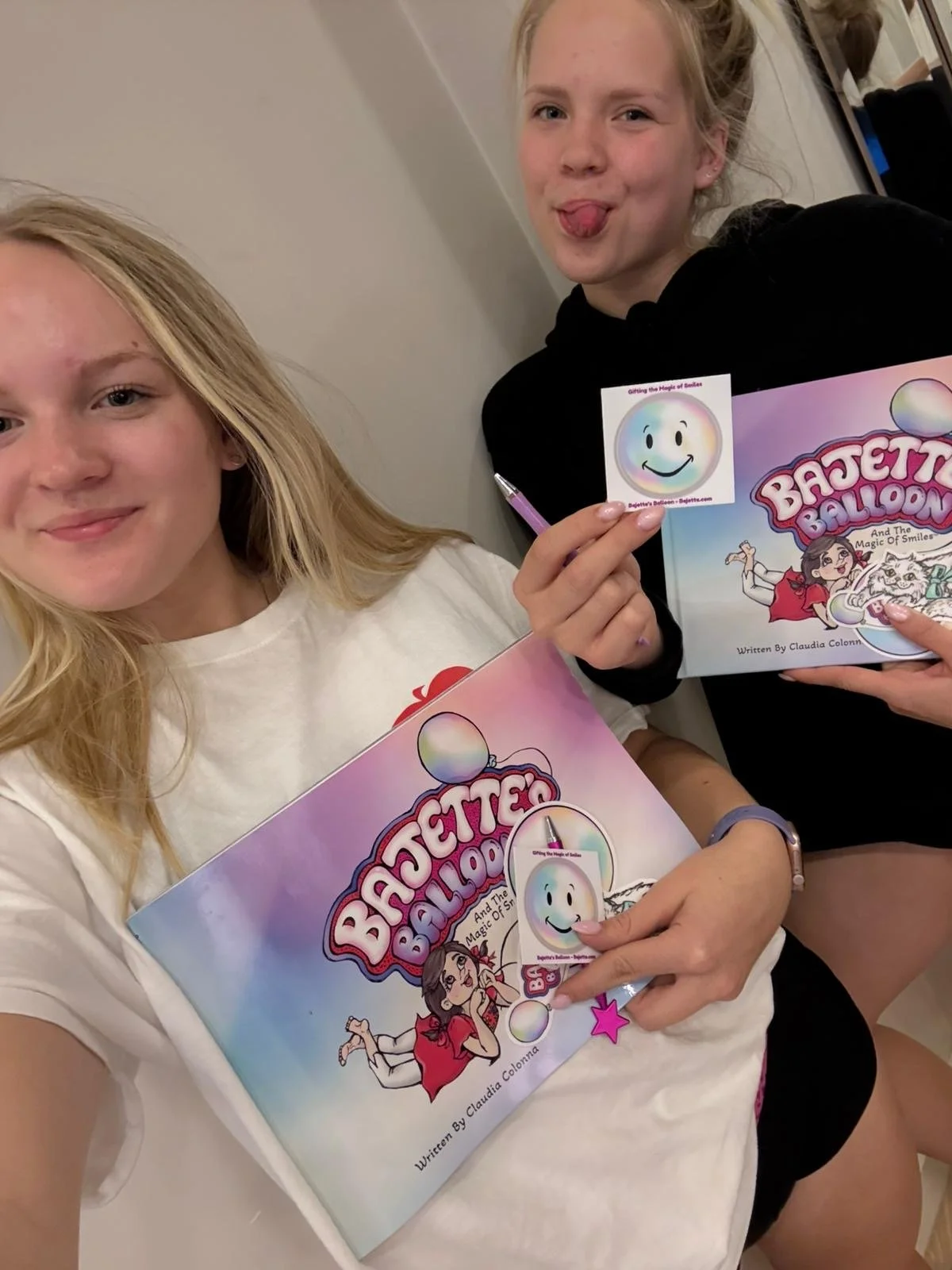 Two young girls holding books titled 'Bachelorette Balloons and The Magic of Smiles,' with one girl also holding a sticker with a happy face and a pink star-shaped charm.