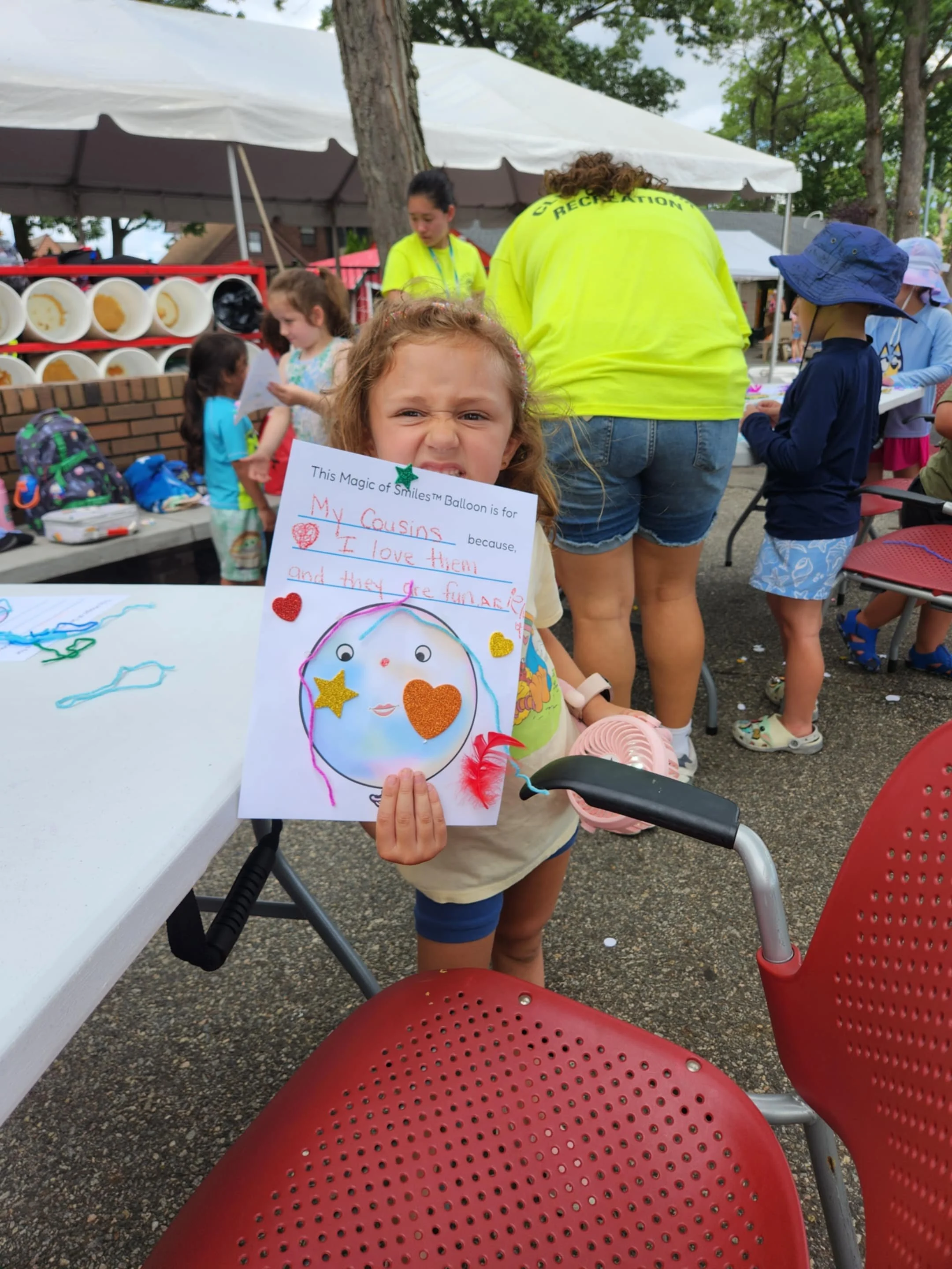 A young girl holding a decorated paper with a face and stars, smiling at a craft fair. Behind her are children and adults at tables, under a tent, with trees and sky in the background.