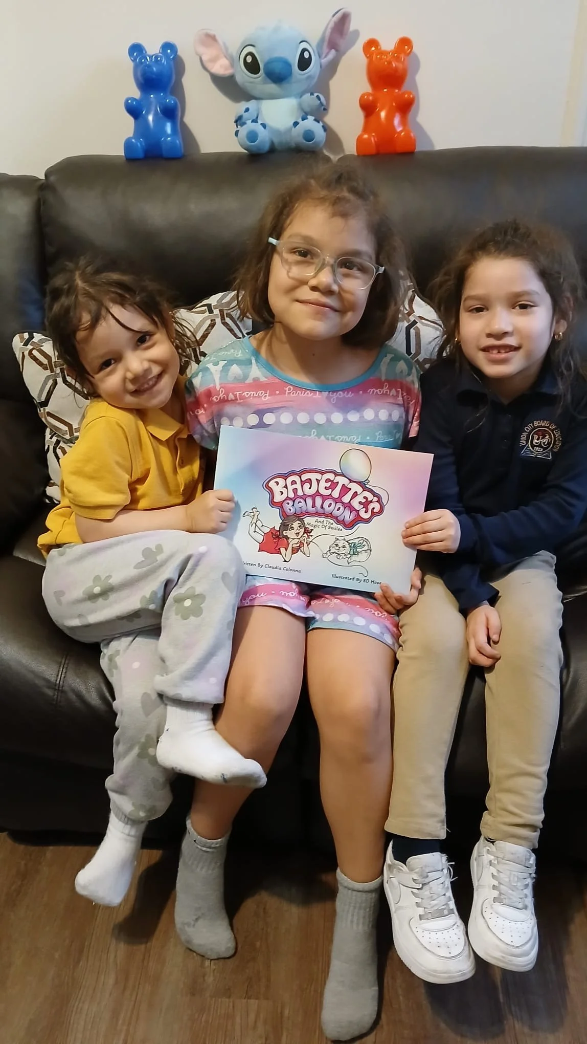 Three young girls sitting on a dark brown leather couch, holding a children's book titled 'Betsy the Balloon'. One girl is in the middle, wearing glasses and colorful pajamas, and the two girls on either side are smiling. Behind them are three stuffe