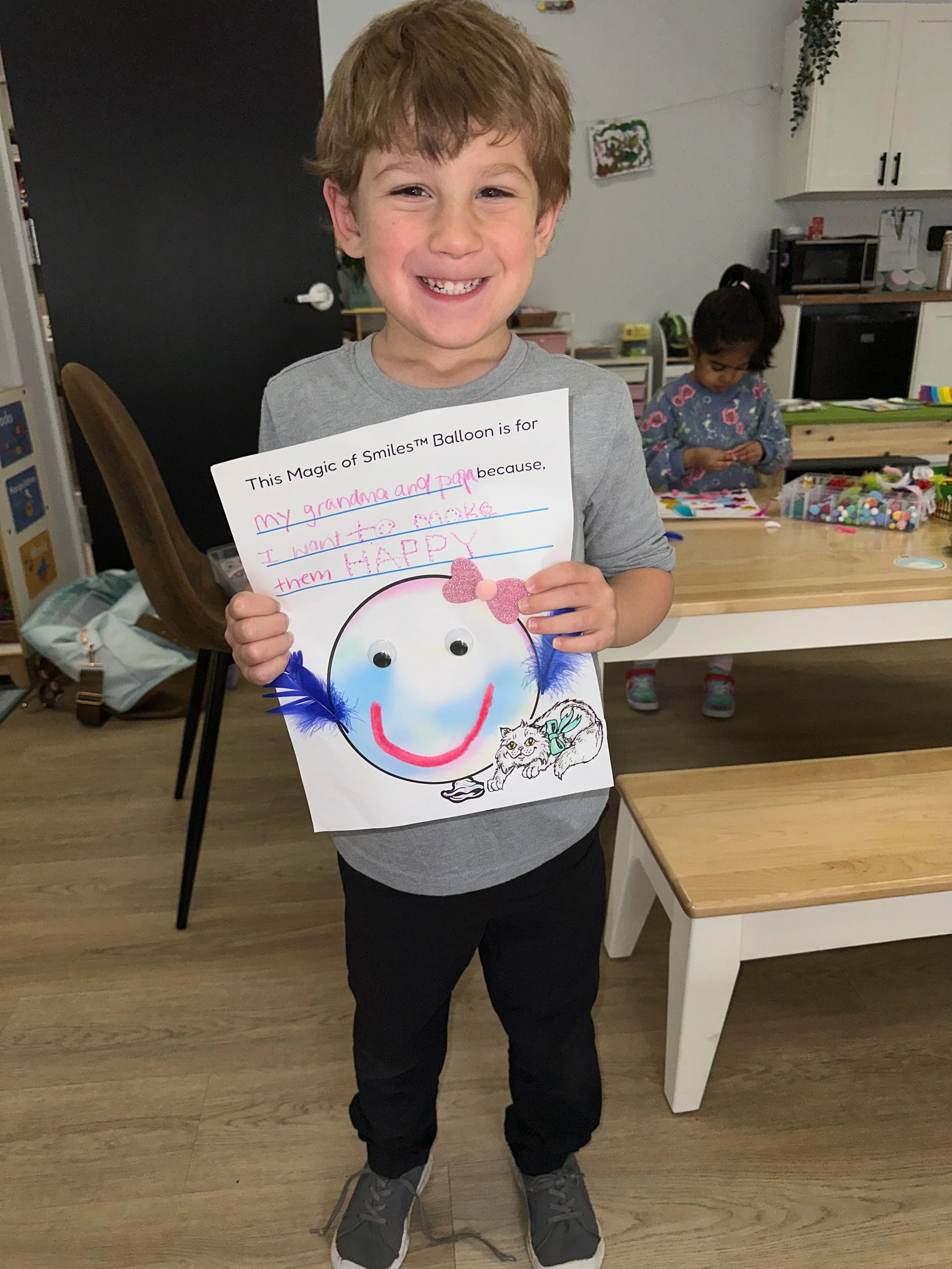 A young boy smiling and holding a craft project with a balloon drawing, googly eyes, feathers, and a pink heart-shaped decoration. In the background, a girl is working at a table with craft supplies.