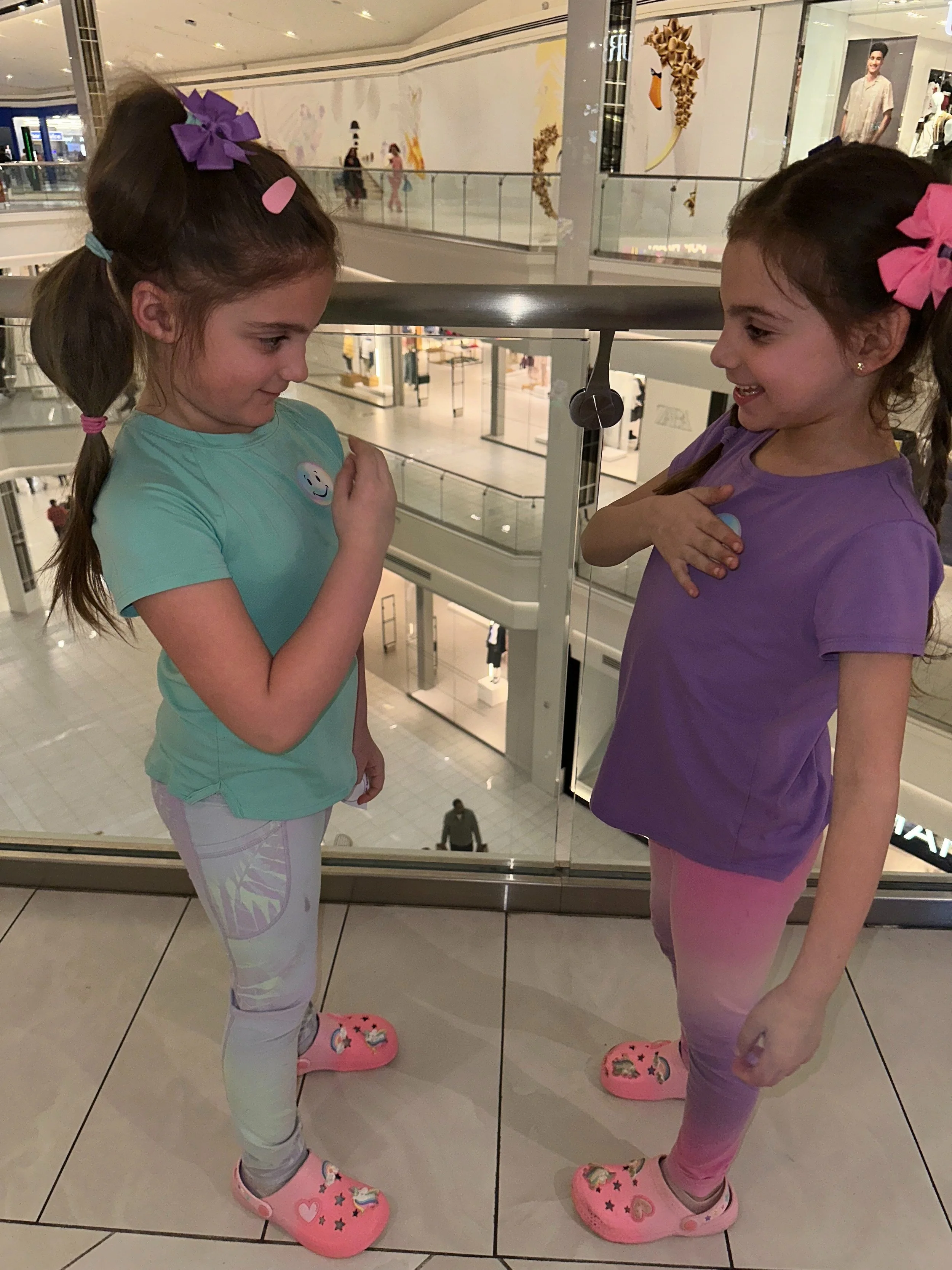 Two young girls are standing in front of a mirror at a shopping mall, smiling and interacting. They are wearing colorful t-shirts, leggings, and pink Crocs, with their hair tied back and decorated with bows and hair clips.