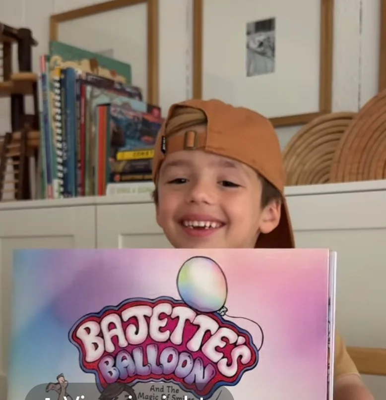 Smiling boy wearing a backwards baseball cap holding a colorful book titled "Bajette's Balloon" in a room with bookshelves and framed pictures.